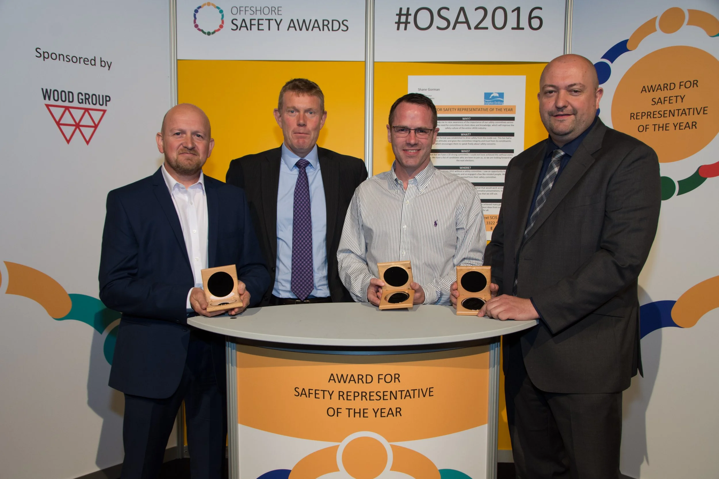 Four men in suits standing at a table holding awards at the Offshore Safety Awards 2016 event. The backdrop displays the event name, hashtags, and sponsors, with the text 'Award for Safety Representative of the Year' visible in the lower part of the 