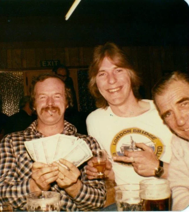 Three men at a bar or pub enjoying drinks and smiling at the camera. The man on the left is holding several dollar bills, the man in the middle is holding a drink, and the man on the right is pointing at the man in the middle. The background has wood