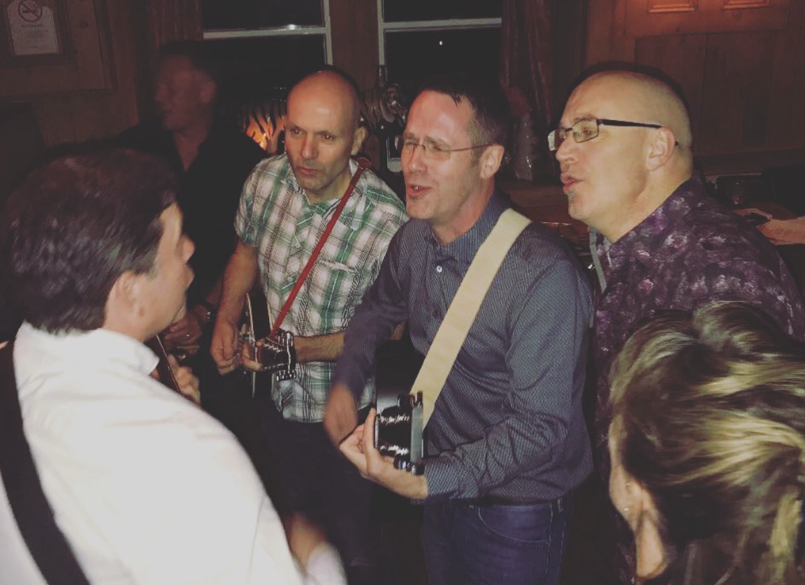 Group of people singing and playing guitars at a social gathering in a cozy, wood-paneled room.