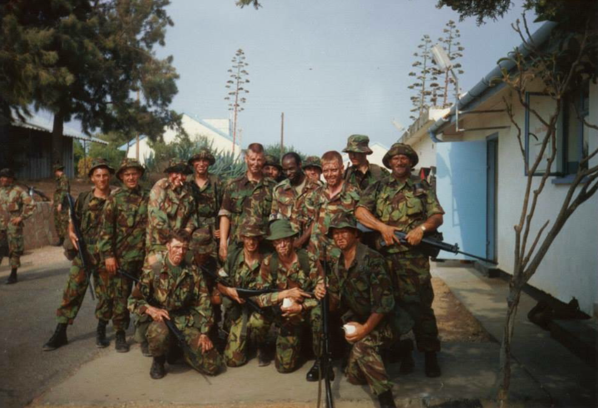 Group of soldiers in camouflage uniforms posing outdoors, some holding rifles, with trees and a building in the background.