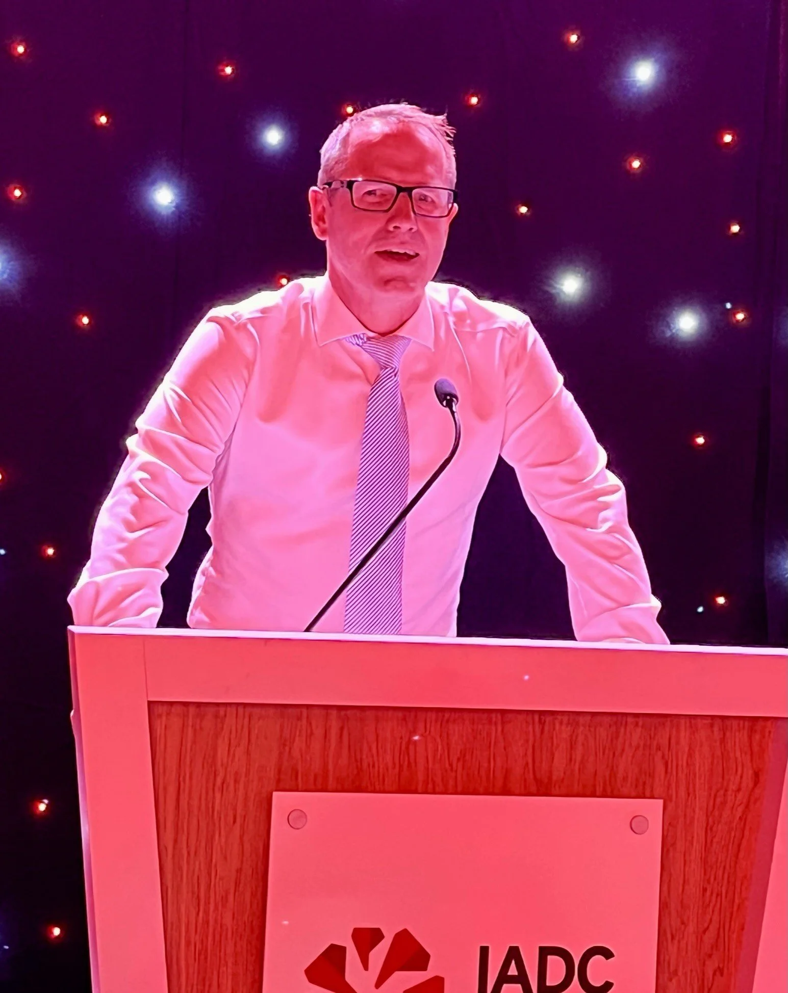 Man with glasses and light-colored shirt and tie standing behind a podium with the IARC logo, in front of a starry background.