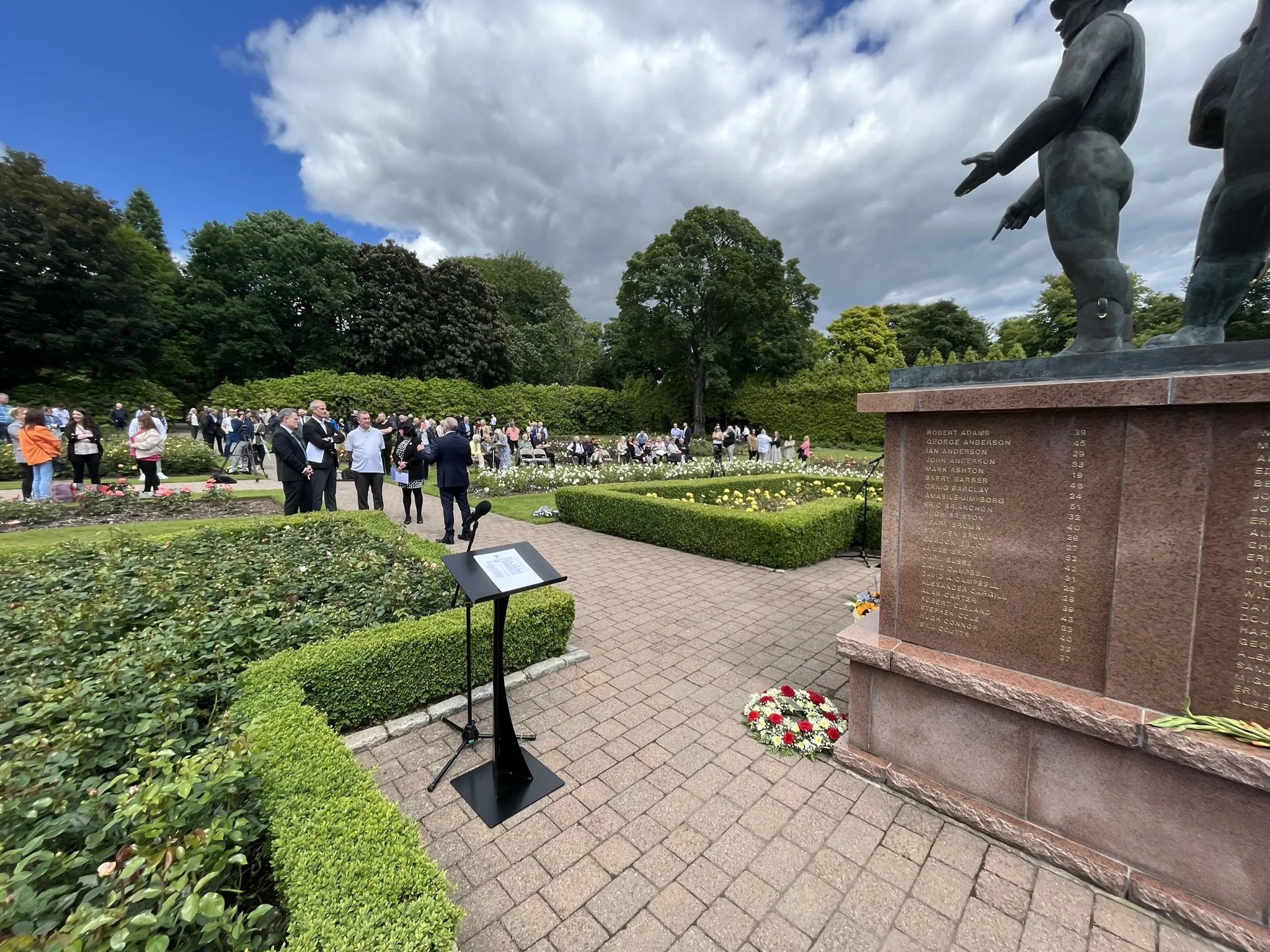 A crowd gathered at a memorial in a park with a large stone monument inscribed with names, surrounded by well-kept bushes, flowers, and trees, under a cloudy sky.