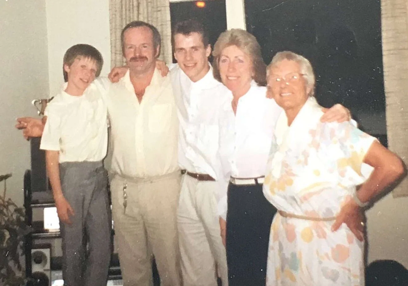 A multi-generational family portrait of six people standing close together indoors, smiling, with one person having his arm around another.