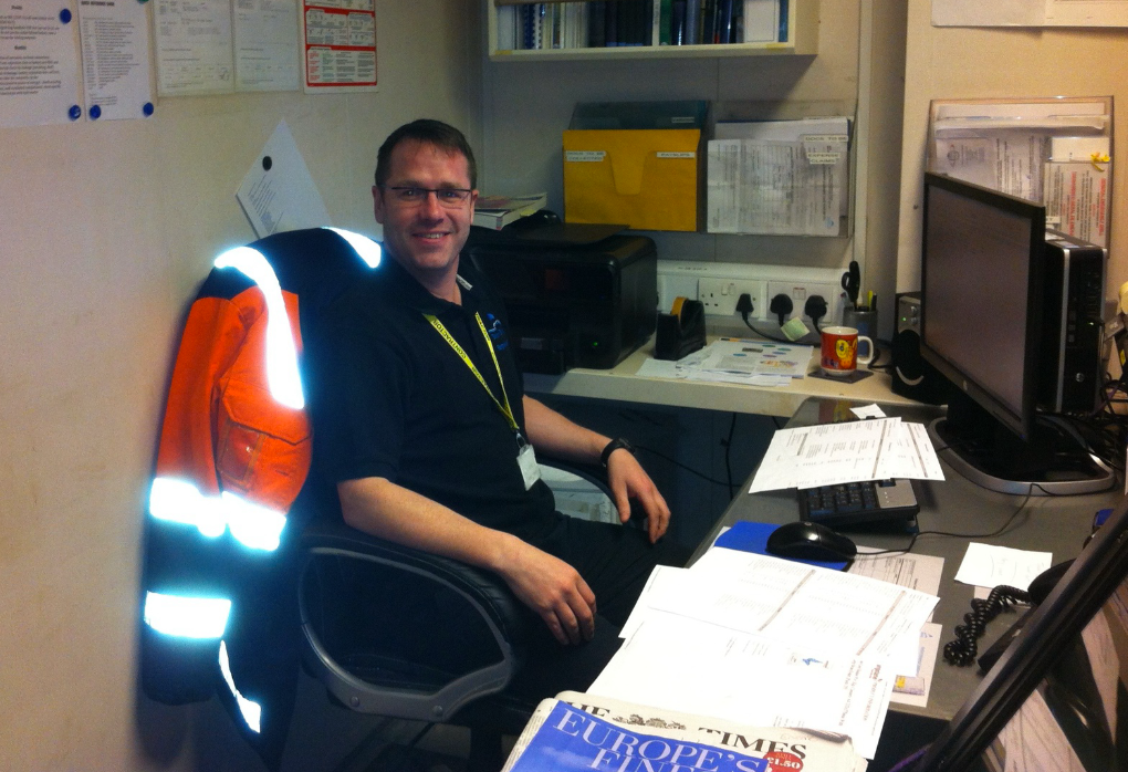 A man sitting at a cluttered desk in an office surrounded by papers, a computer, and office supplies, wearing a dark uniform and a high-visibility reflective safety vest hung on the back of his chair, smiling at the camera.