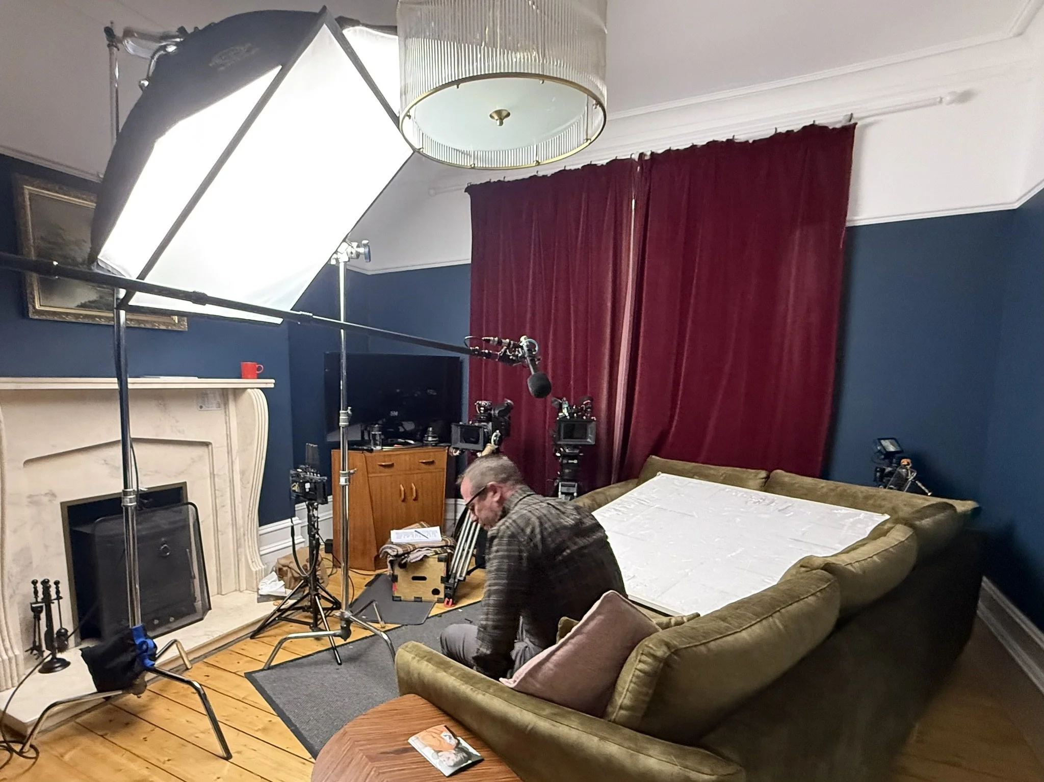 A behind-the-scenes of a film or photography set in a living room, including lighting equipment, cameras, a man adjusting gear, a vintage fireplace, a marble coffee table, a green velvet sofa, red curtains, and a man with glasses sitting on the floor