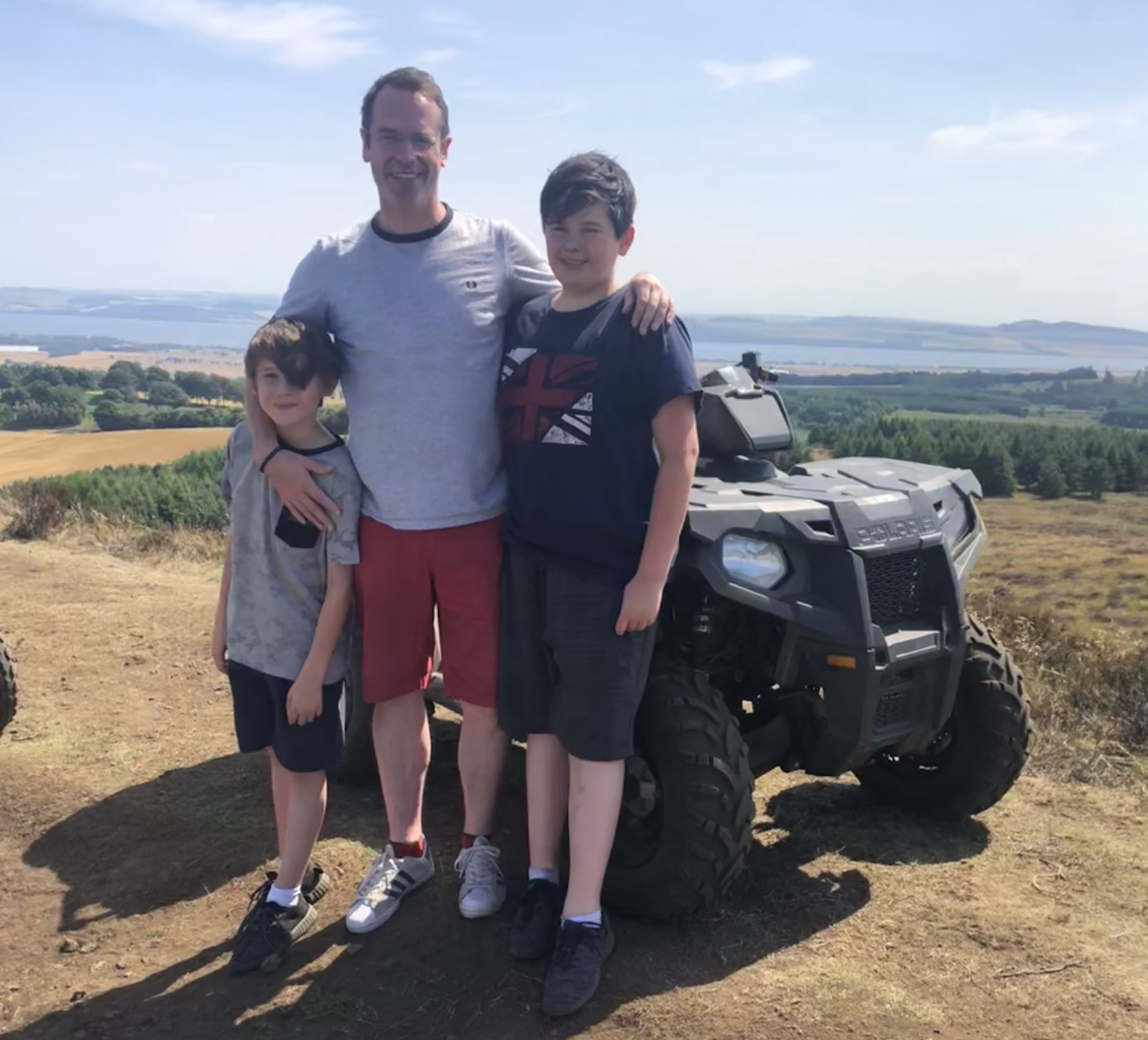 A man and two boys standing outdoors on a hilltop with a Polaris off-road vehicle and a scenic landscape in the background.