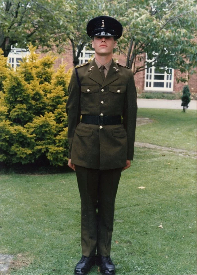 A person in a military uniform standing outdoors on grass with trees and a building in the background.