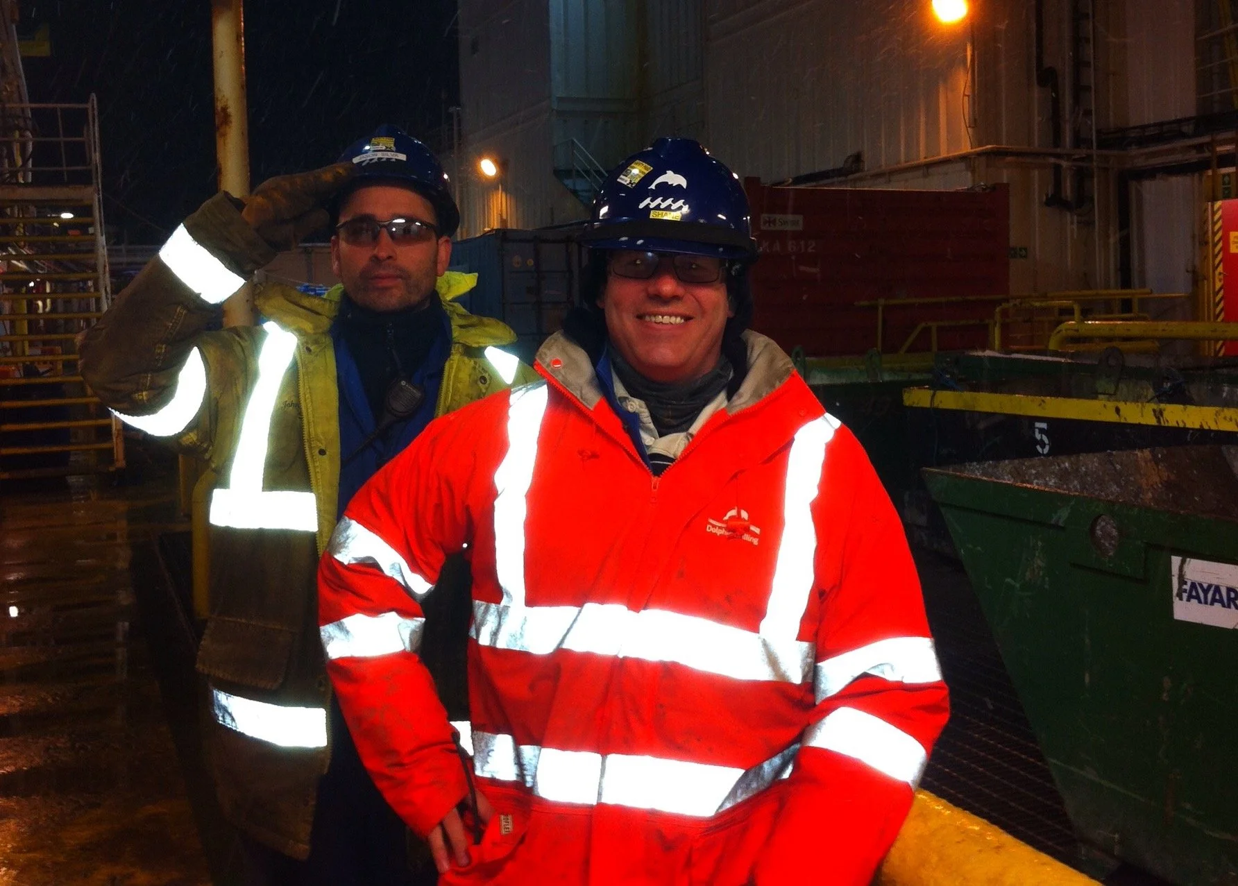 Two construction workers standing outdoors at night in rainy weather, wearing reflective gear and helmets. One is in a yellow jacket with reflective stripes, the other in an orange jacket with reflective stripes, both smiling and posing for the photo