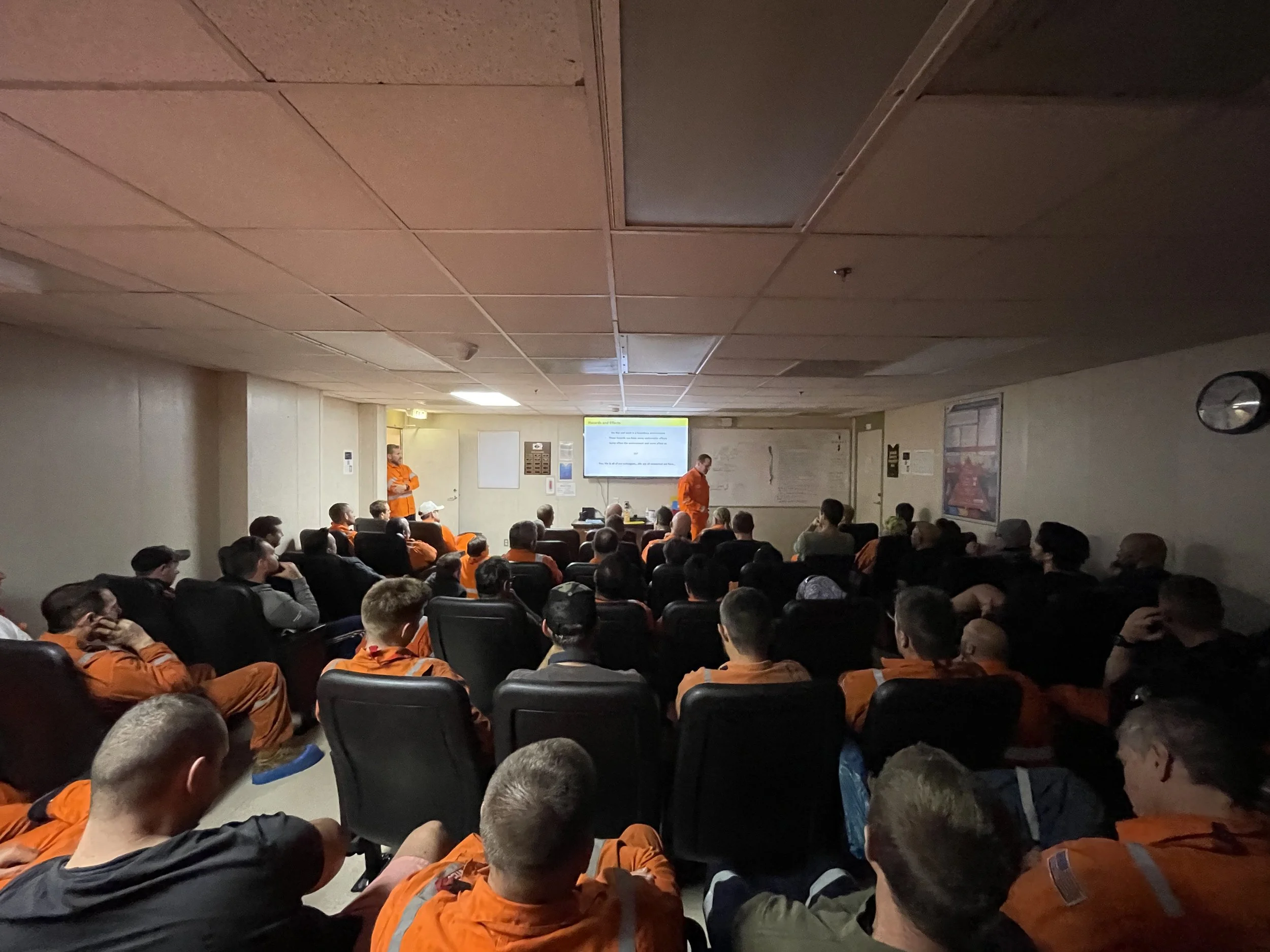 A group of people, many in orange uniforms registered for a presentation or meeting in a room with a ceiling-mounted screen displaying a slide.