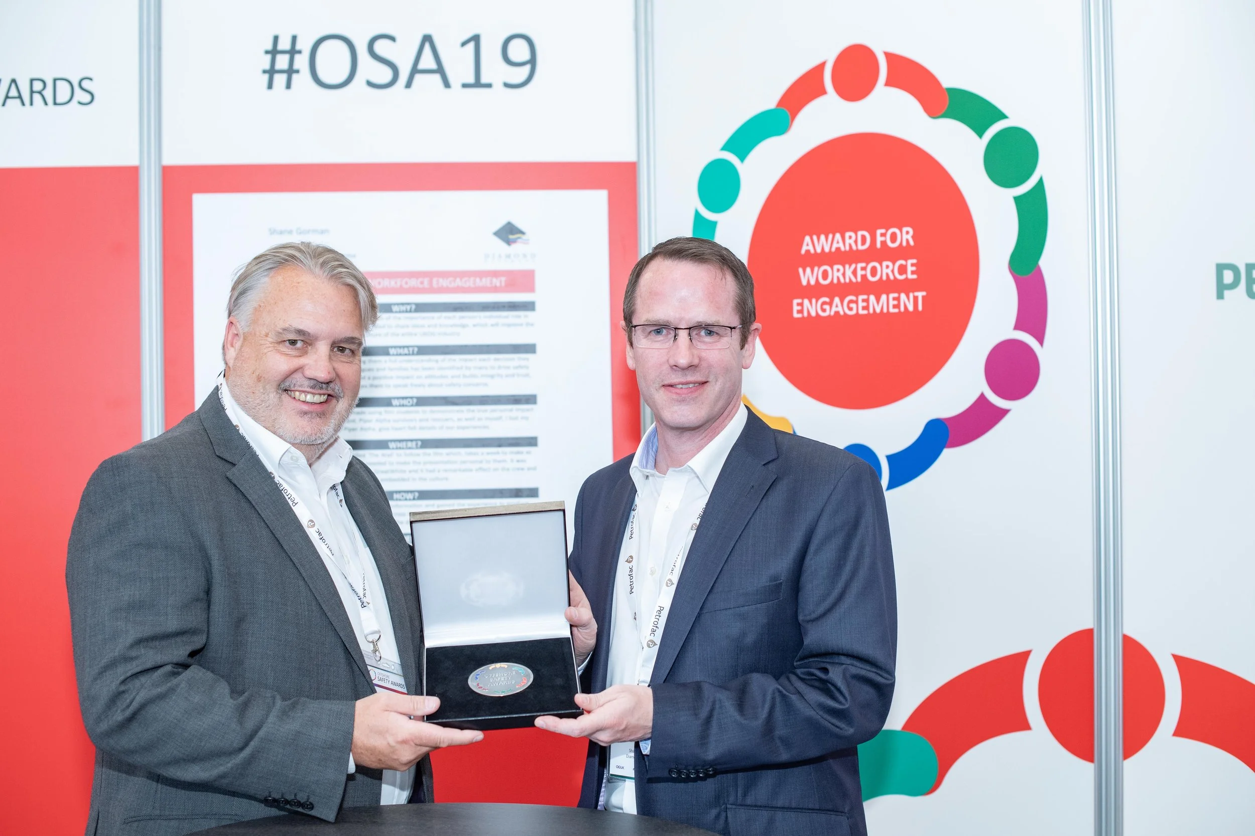 Two men wearing suits and glasses are standing together at a conference, holding a framed award. Behind them are colorful logos and signs related to workforce engagement and a hashtag #OSA19.