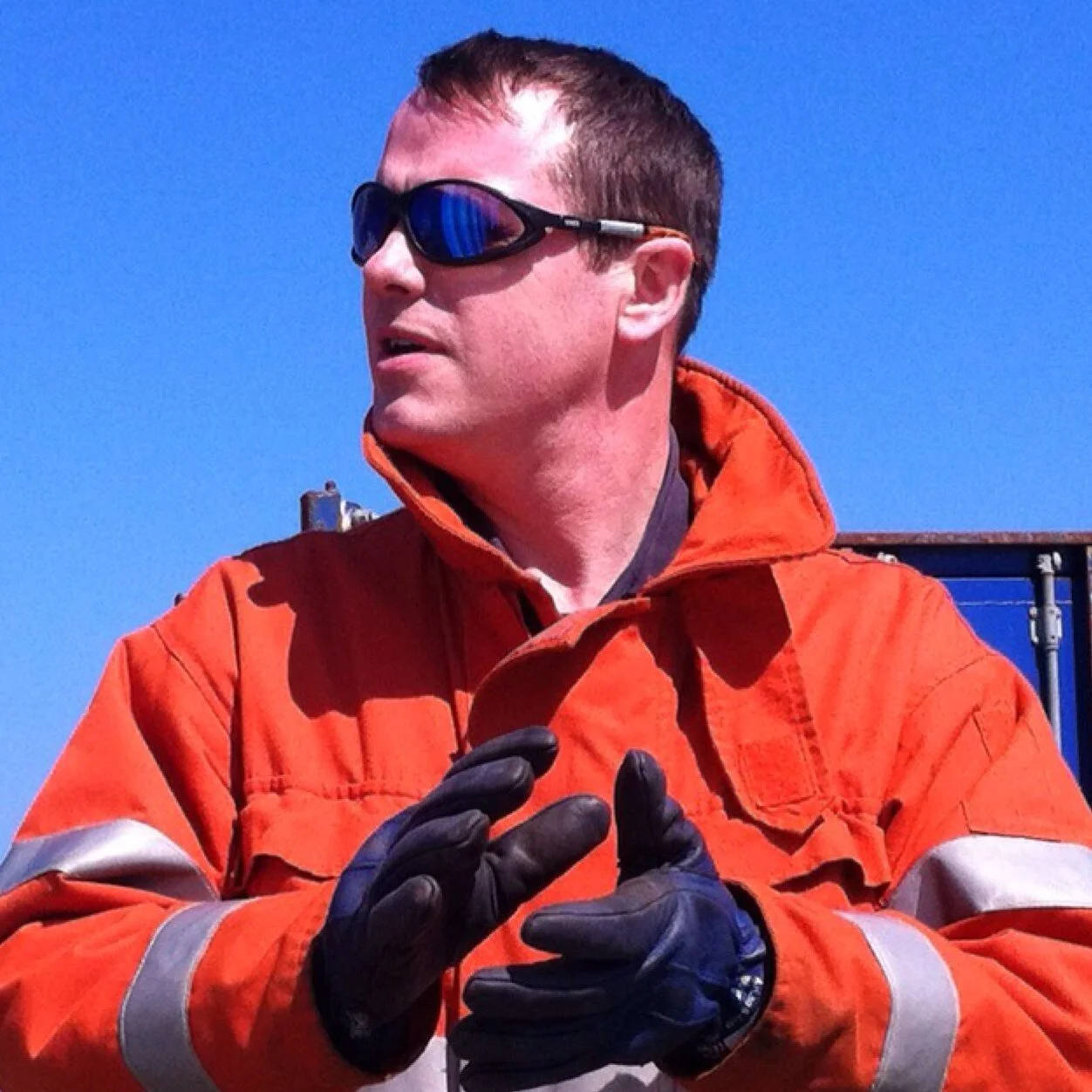 Man wearing orange safety jacket, sunglasses, and gloves, standing outdoors against a clear blue sky.
