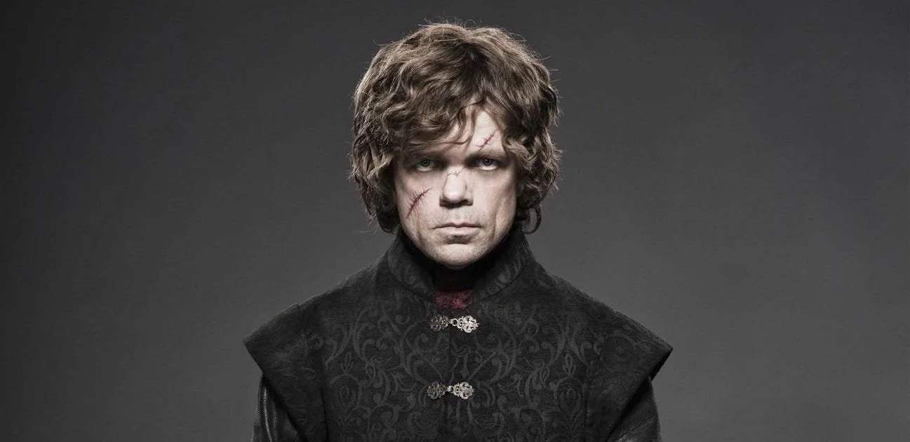 A man with curly hair, facial scars, and a serious expression wearing a dark, ornate jacket against a plain gray background.