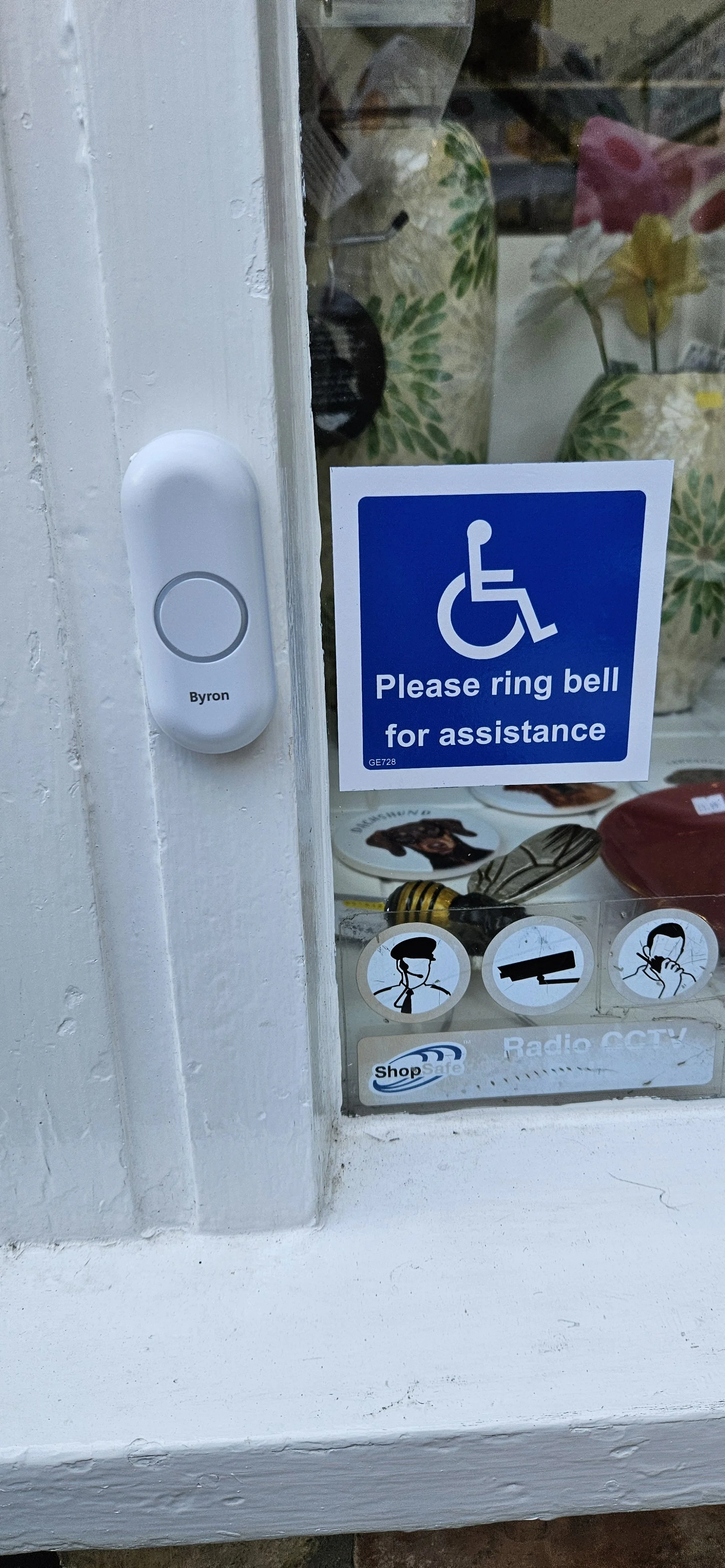 A window with a blue sign that has a wheelchair symbol and the text 'Please ring bell for assistance.' There is a white device labeled 'Byron' mounted on the side of the window, and various decorative items and stickers are visible inside the window.