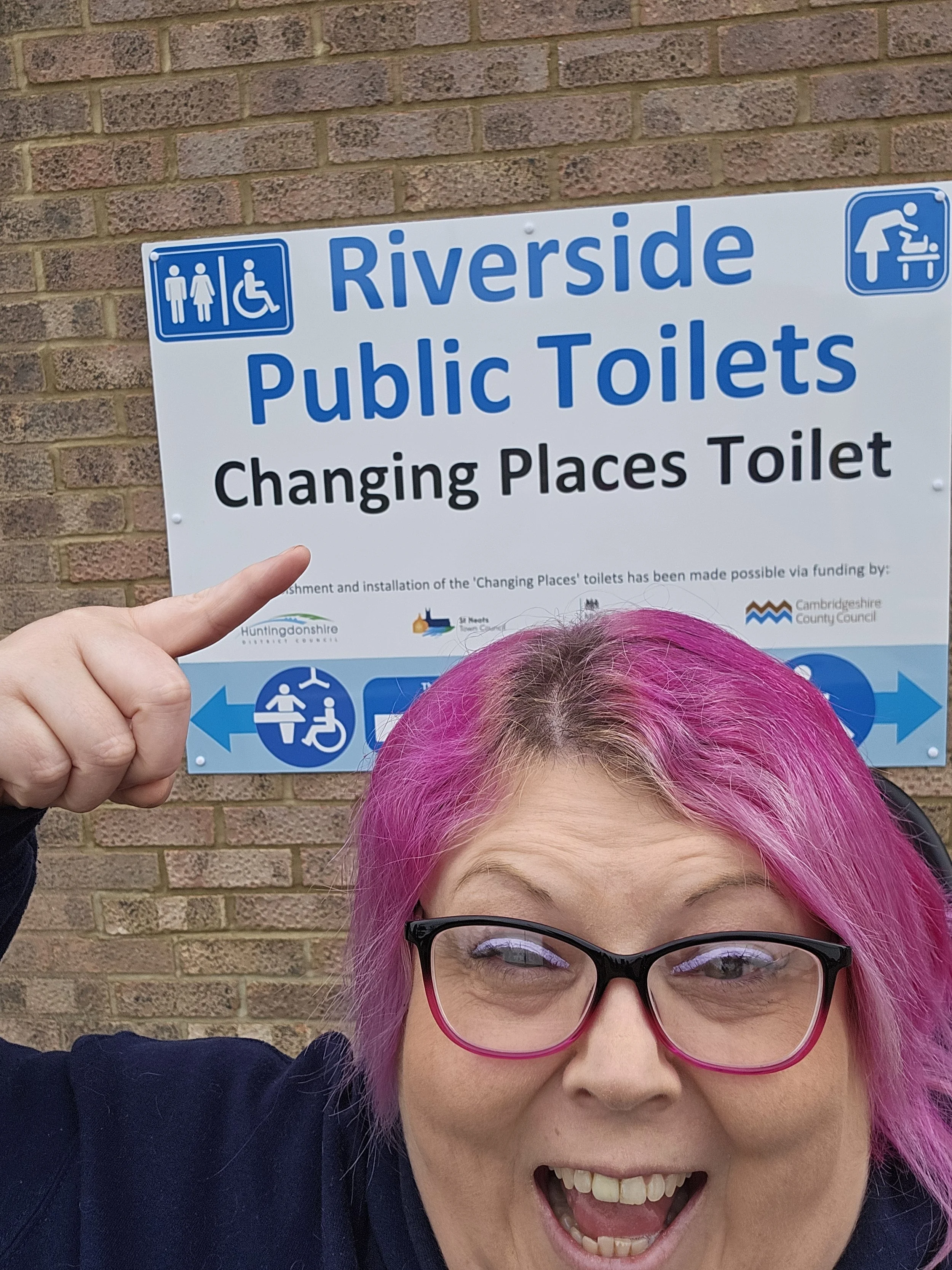 Woman with colorful pink hair and glasses smiling in front of a brick wall, pointing at a sign for Riverside Public Toilets and Changing Places Toilet.