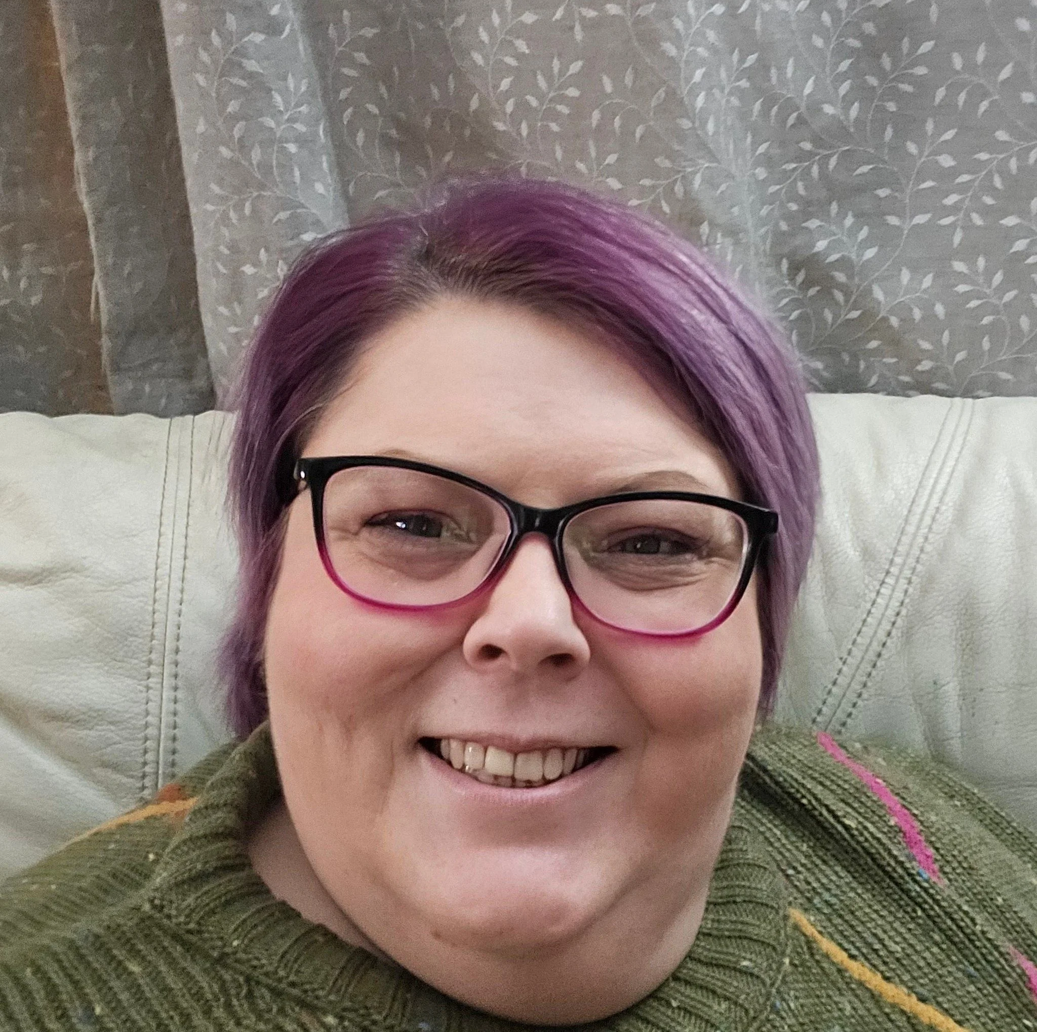 A smiling woman with purple hair, wearing black glasses, lying on a bed with a patterned green blanket and a gray wallpaper with a white leaf pattern in the background.