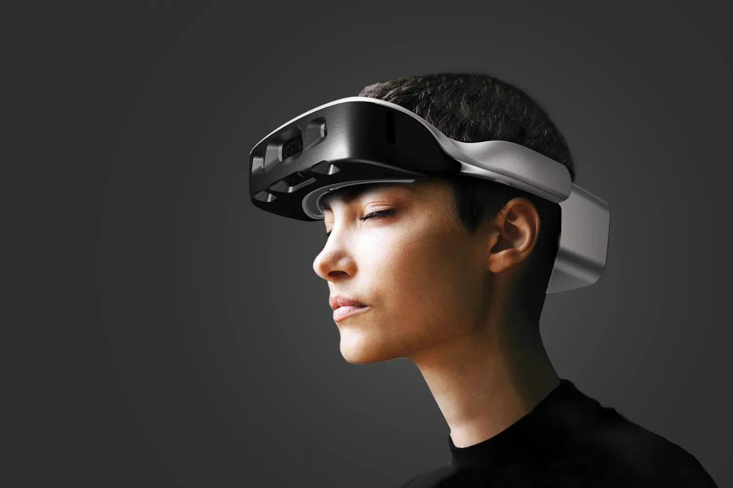 Person with short dark hair wearing a virtual reality headset with their eyes closed against a dark gray background.