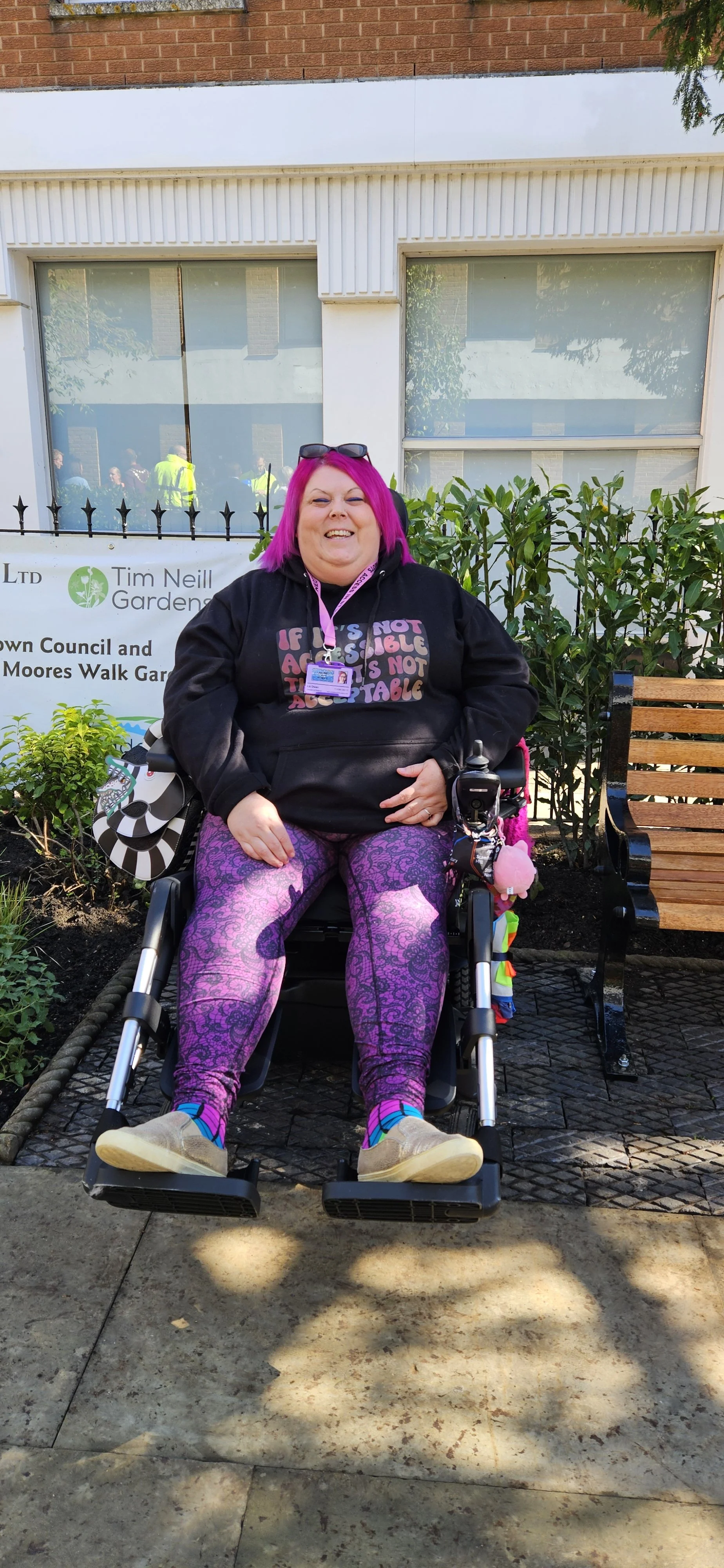 Me (pink hair, Access in St Neots hoodie, purple leggings, gold shoes) in my wheelchair parked in one of the 2 new spaces for wheelchair users
