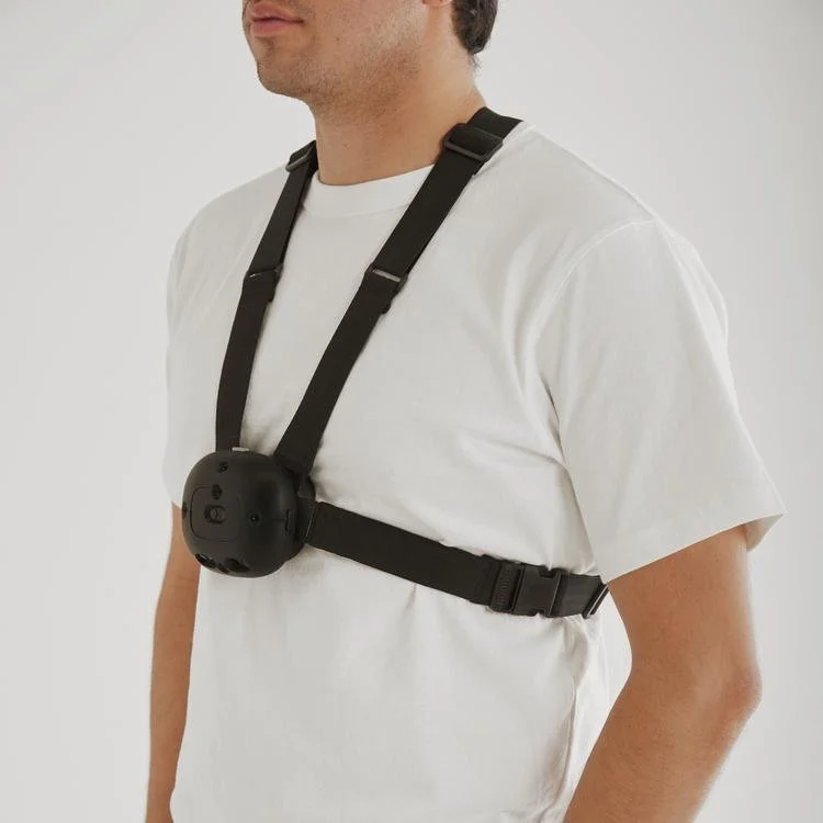A man wearing a white T-shirt and a black chest strap with a round, black electronic device attached.