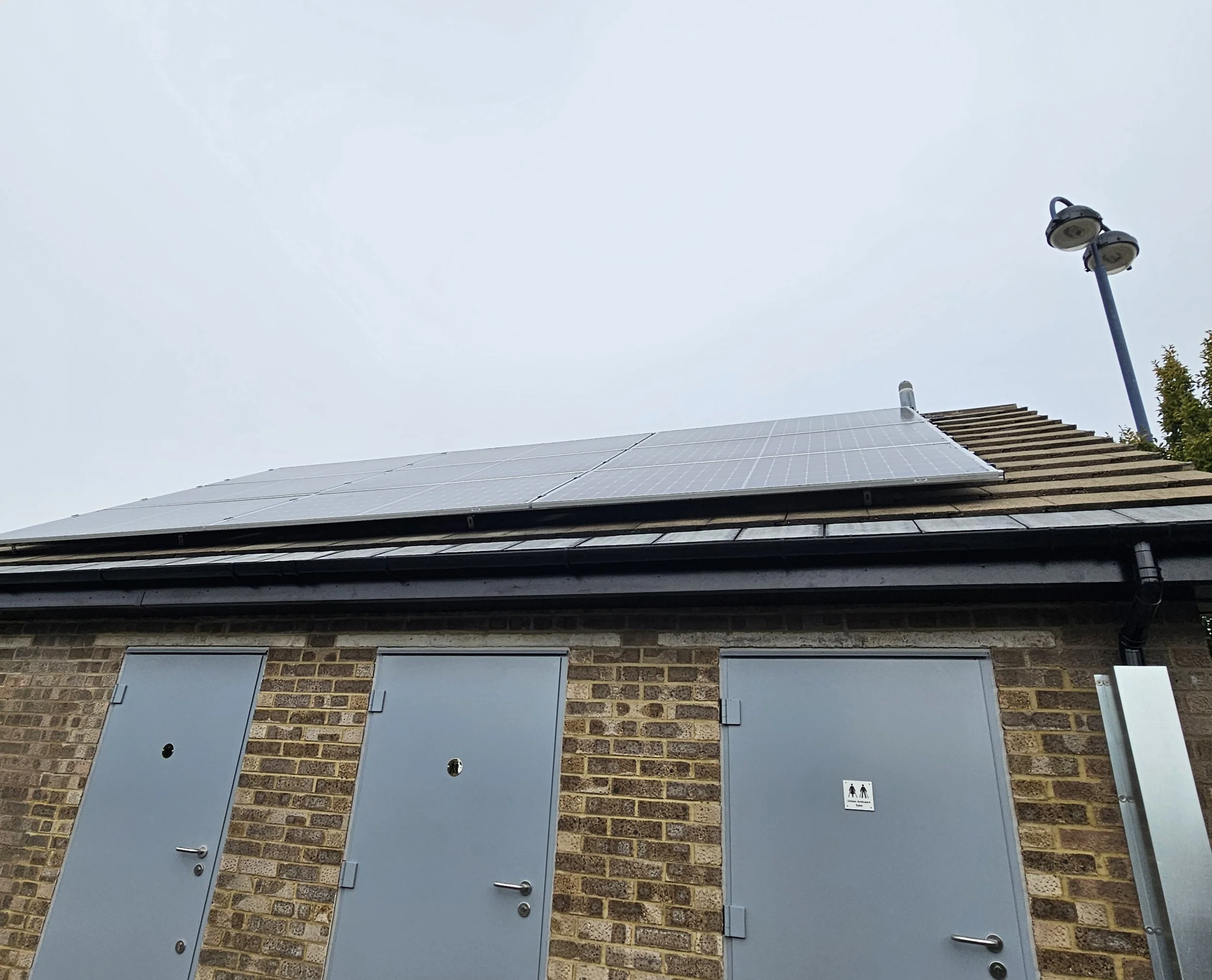 Solar panels installed on a residential roof with a brick facade, two grey utility doors with blue shutters, and a small sign indicating bathroom facilities.