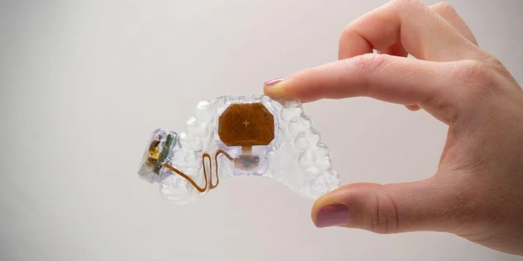 A person's hand holding a clear dental device or retainer with a small electronic component attached inside, featuring a brown battery.