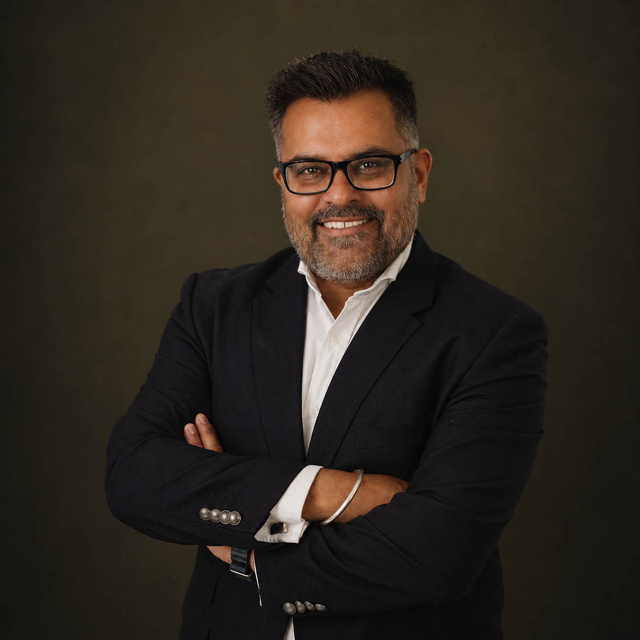 A well-dressed man with glasses, a beard, and gray hair, smiling with arms crossed, wearing a black blazer and white shirt, against a dark background.