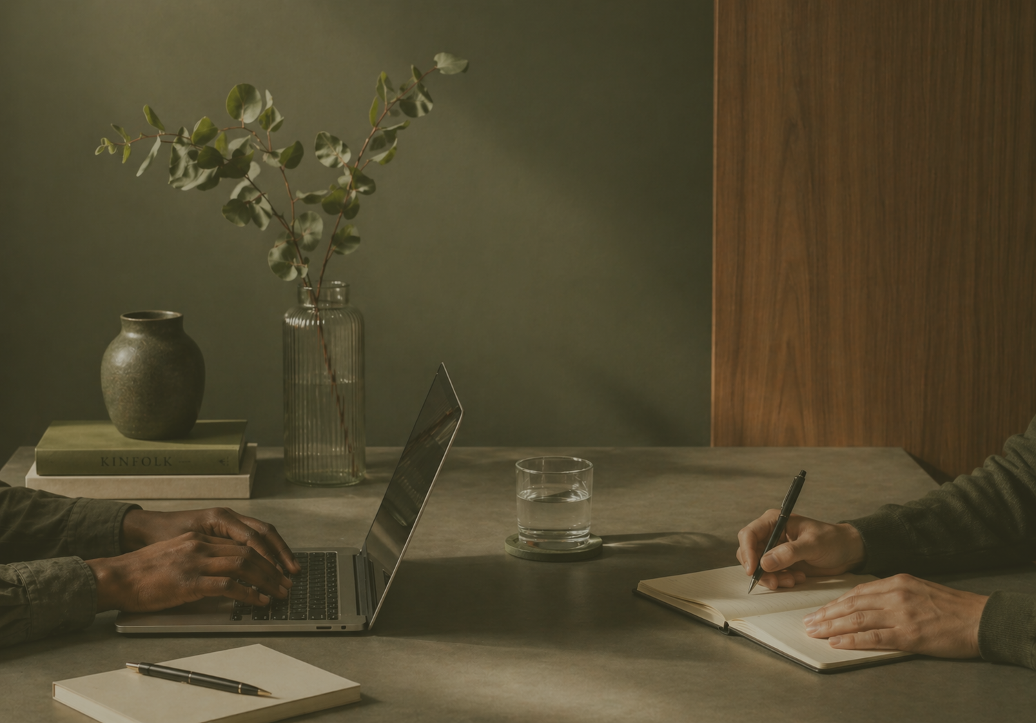 A workspace with two people: one typing on a laptop, the other writing in a notebook. A glass of water, a green book titled 'KINFOLK,' a ceramic vase, and a tall glass vase with green leaves are on the table. The background has a green wall and wooden panel.