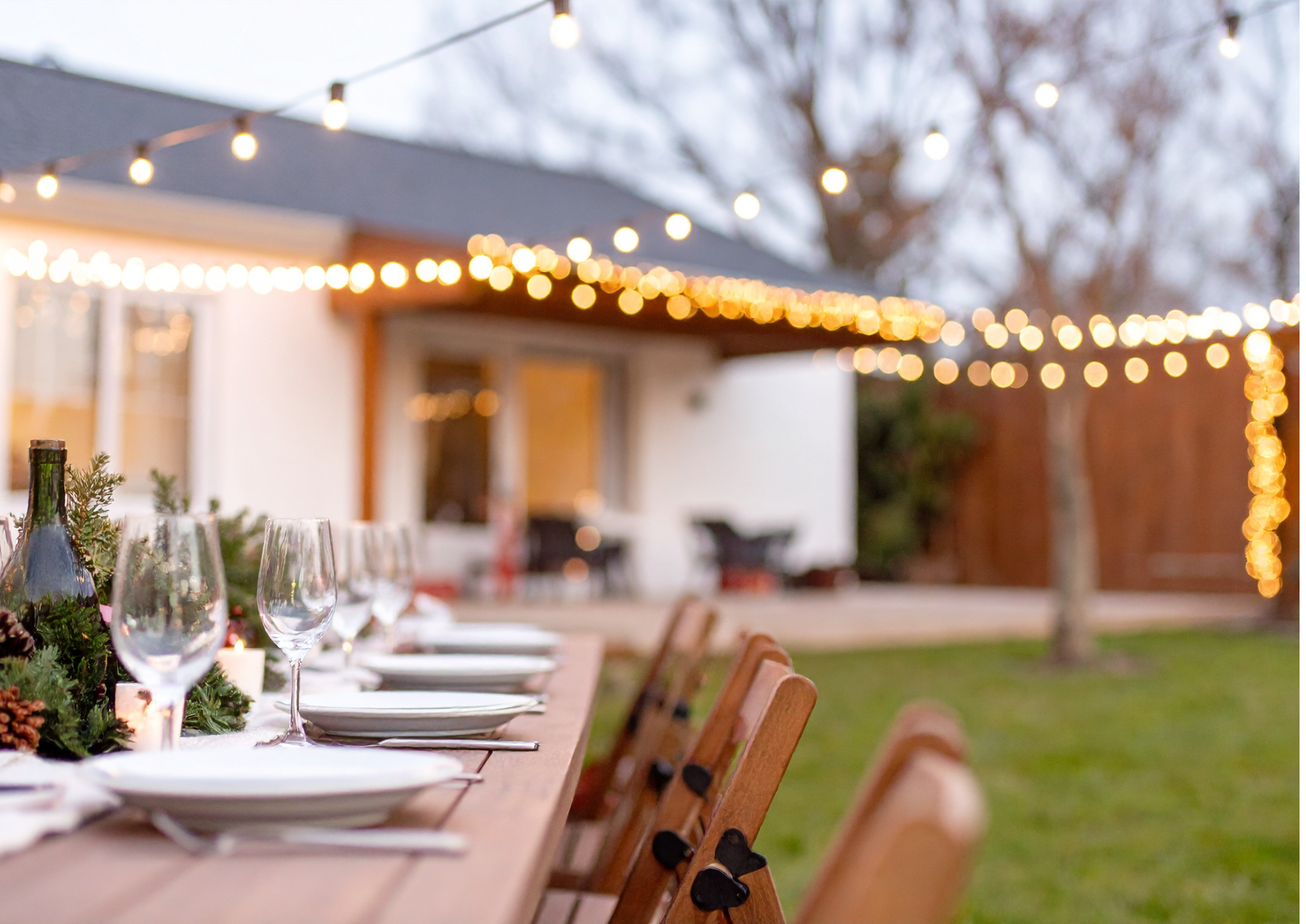 A long outdoor dining table set for an event with wine glasses, plates, and greenery, illuminated by string lights at dusk or evening.