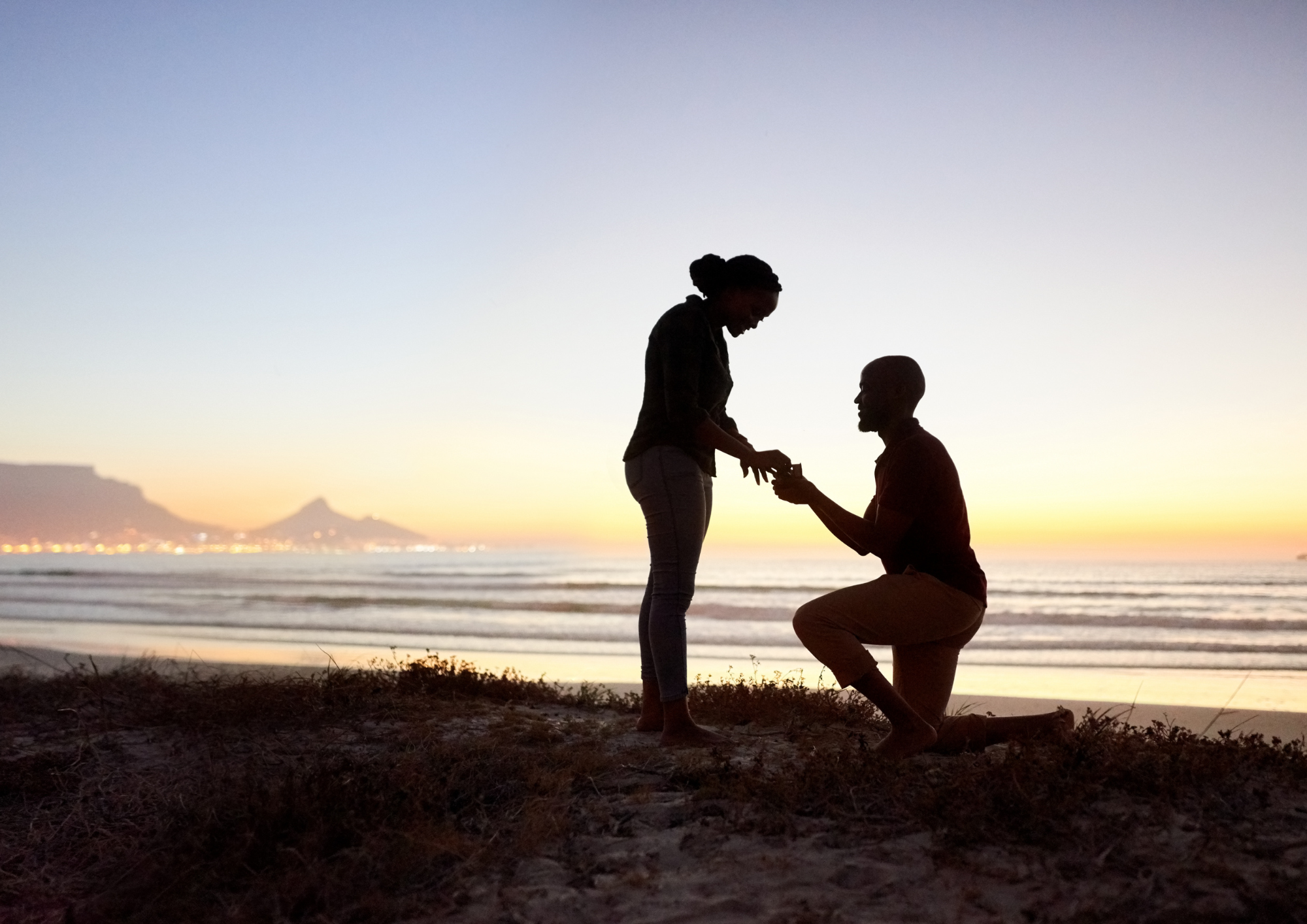 A silhouette of a man kneeling on one knee proposing to a woman on the beach during sunset, with a city and mountain in the background.