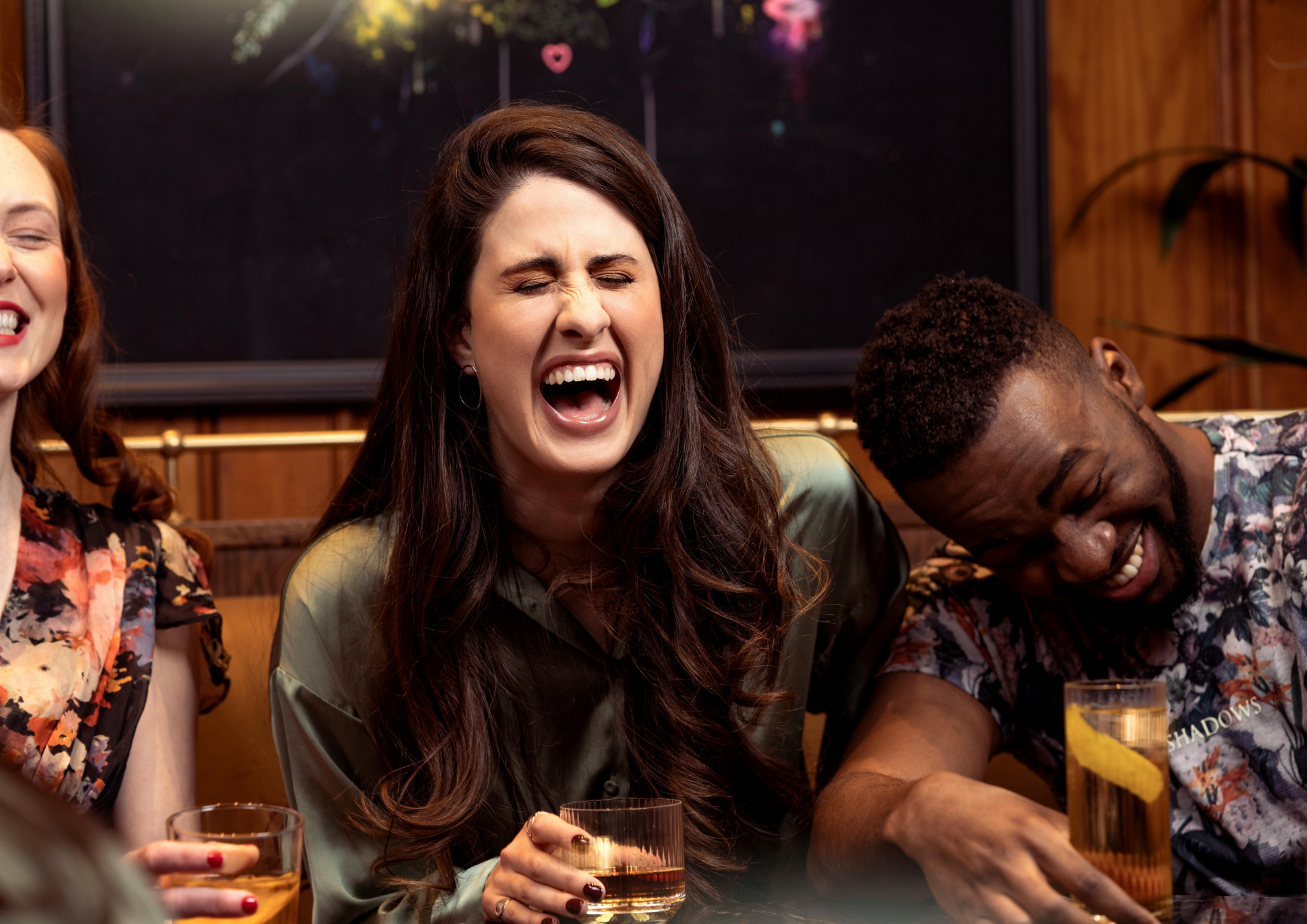 Three people at a social gathering laughing and enjoying drinks in a cozy, warmly-lit room. A woman with long brown hair in the center is closing her eyes and laughing, holding a glass of golden drink. To her right is a man with short curly hair, also laughing and leaning forward. Partially visible on the left is a woman with red hair. The background features a black chalkboard and wooden paneling.