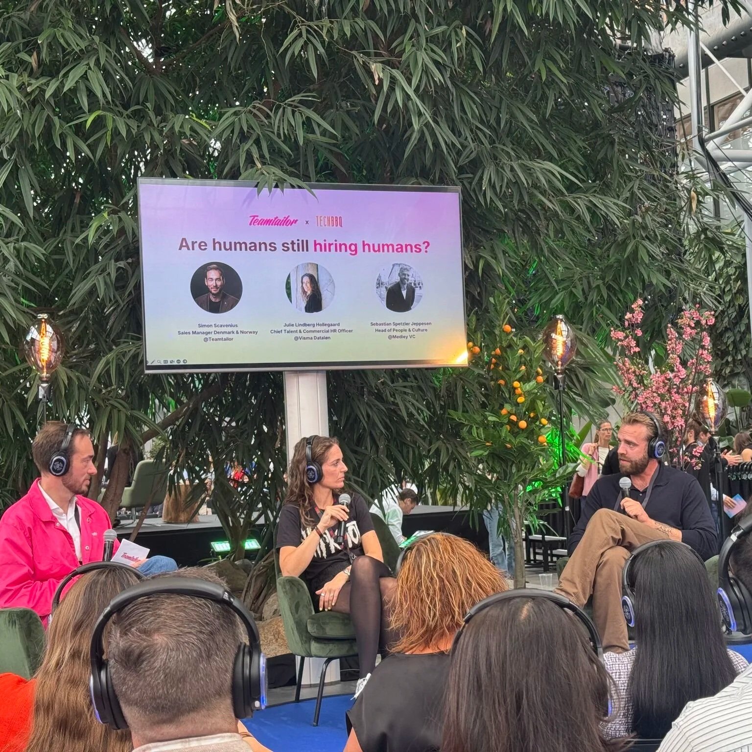 Paneldiskussion med tre personer, der taler i mikrofoner, på en scene omgivet af grønne planter. En stor skærm over diskussionen viser emnet 'Are humans still hiring humans?' med billeder og titler af paneldeltagerne.