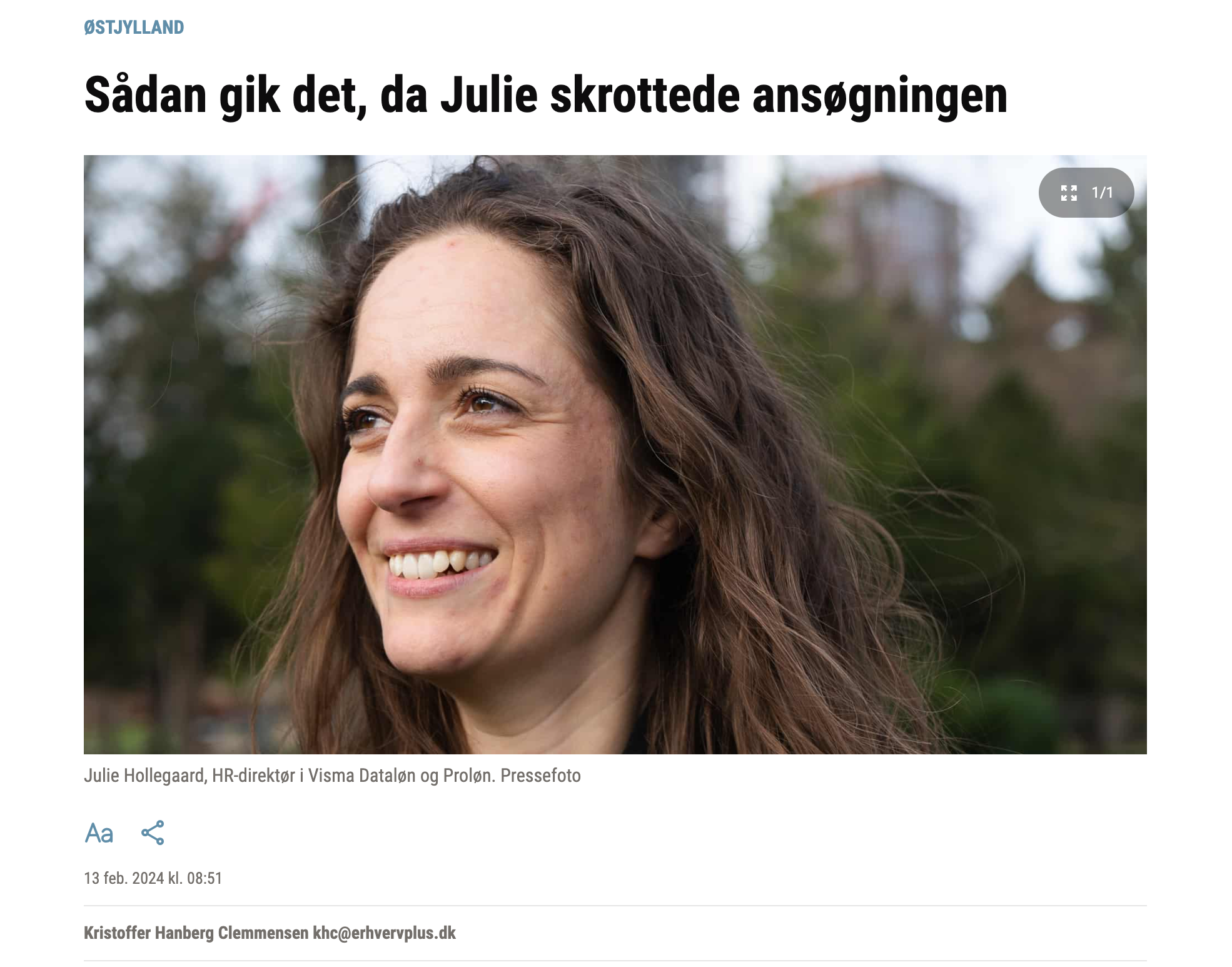Et nærbillede af en smilende kvinde med brunt, lang, bølget hår, set ude i en park med blade og træer i baggrunden.