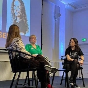 Tre kvinder sidder i en paneldiskussion foran en stor skærm, der viser et billed af en kvinde.