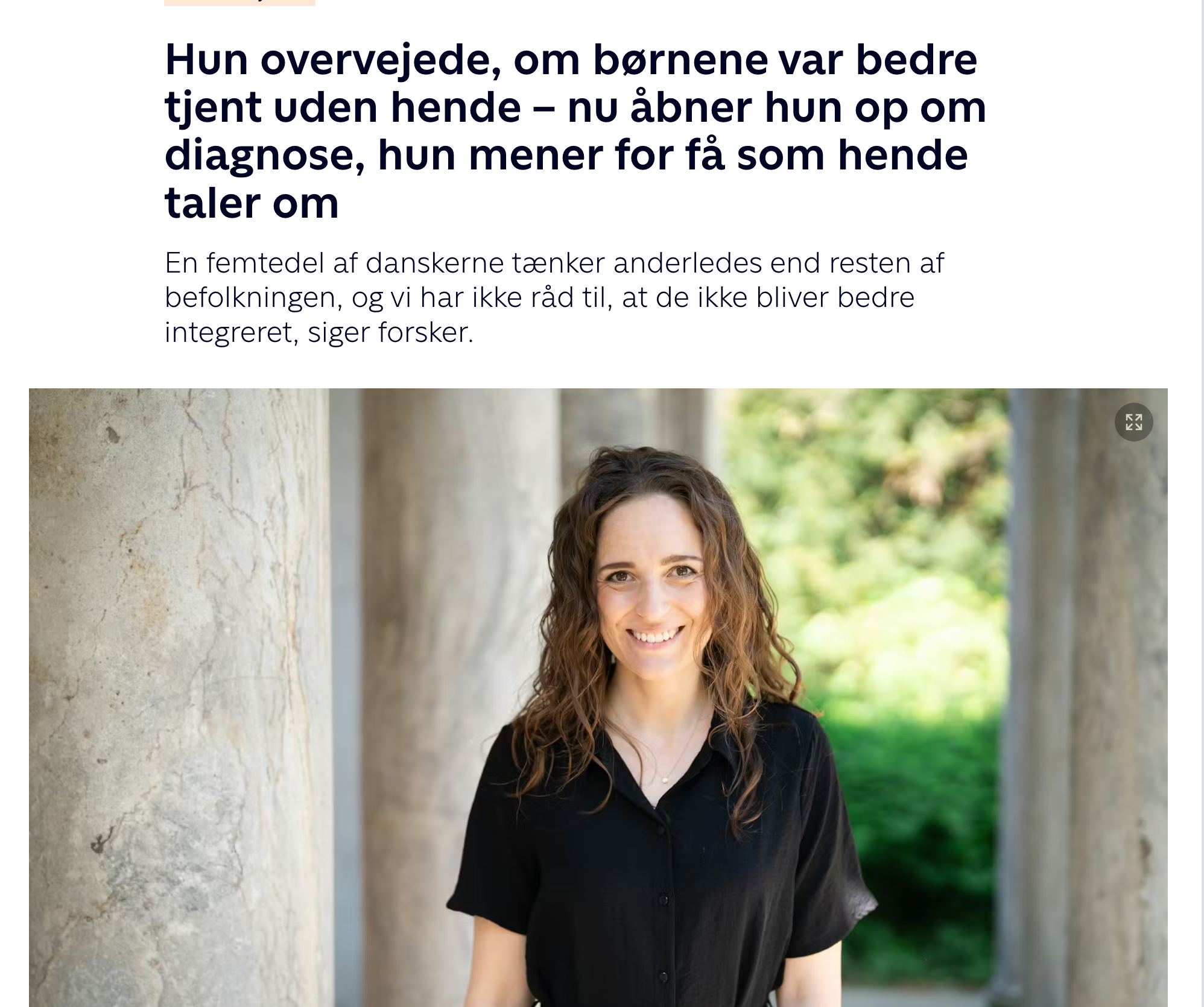 Glad kvinde med brunt, krøllet hår stående udendørs ved en væg, med grønne træer i baggrunden.