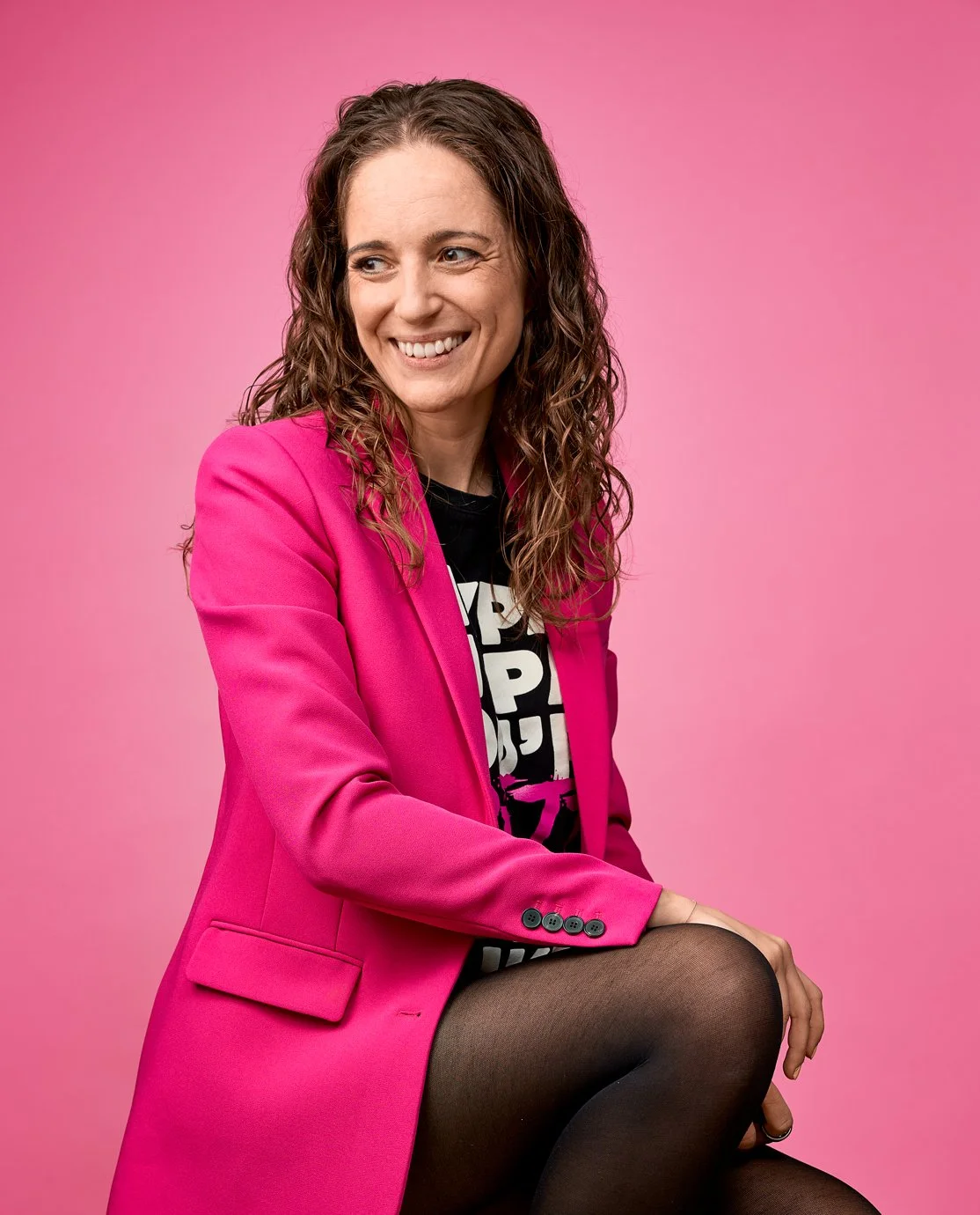 En kvinde med krøllet brunt hår iført en pink blazer og sort tøj, sidder imod en rosa baggrund og smiler.