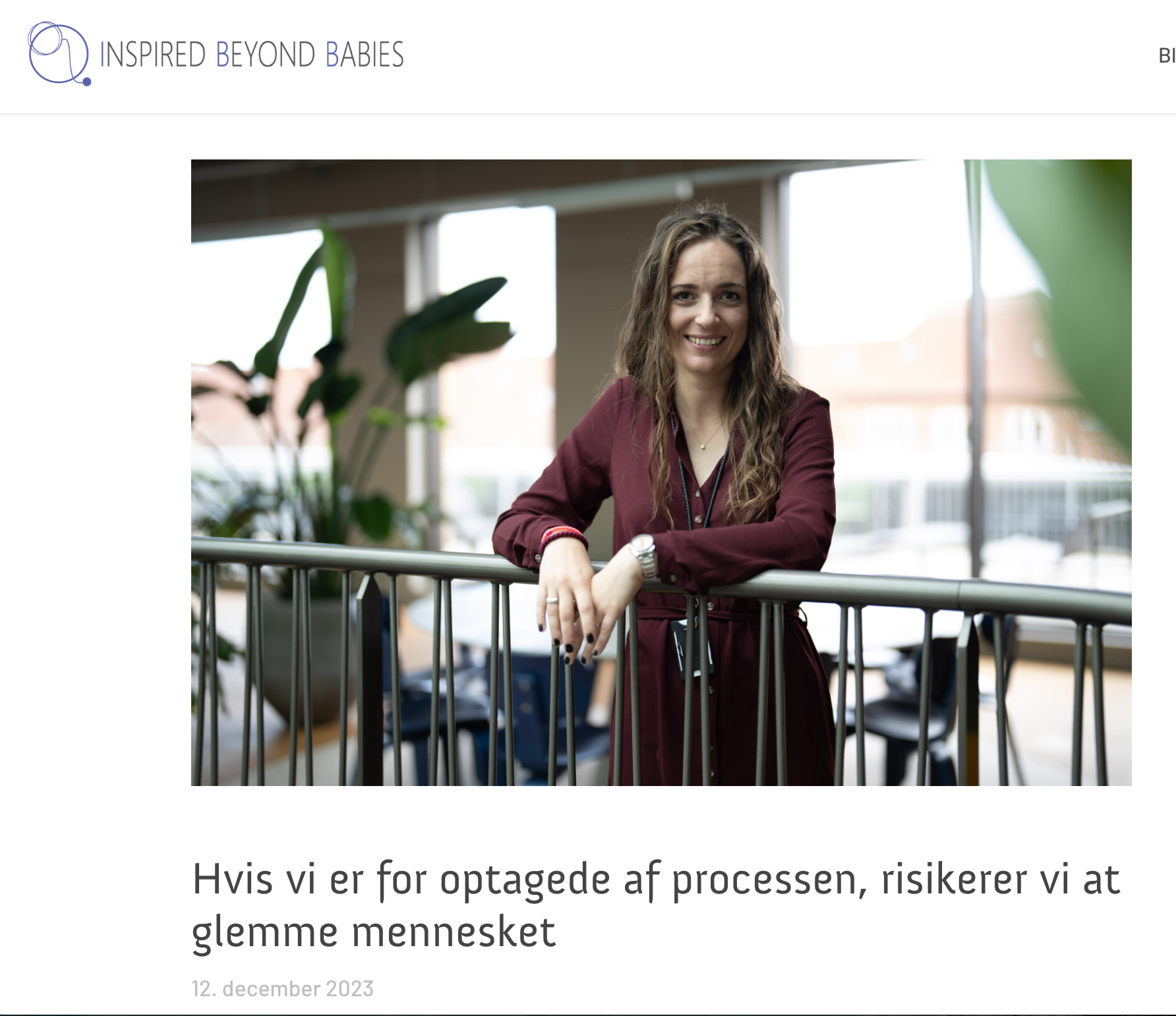 Billede af en smilende kvinde med brunt, bølget hår, iført en vinrød kjole, der står foran store vinduer, omgivet af grønne planter i et indendørs miljø.