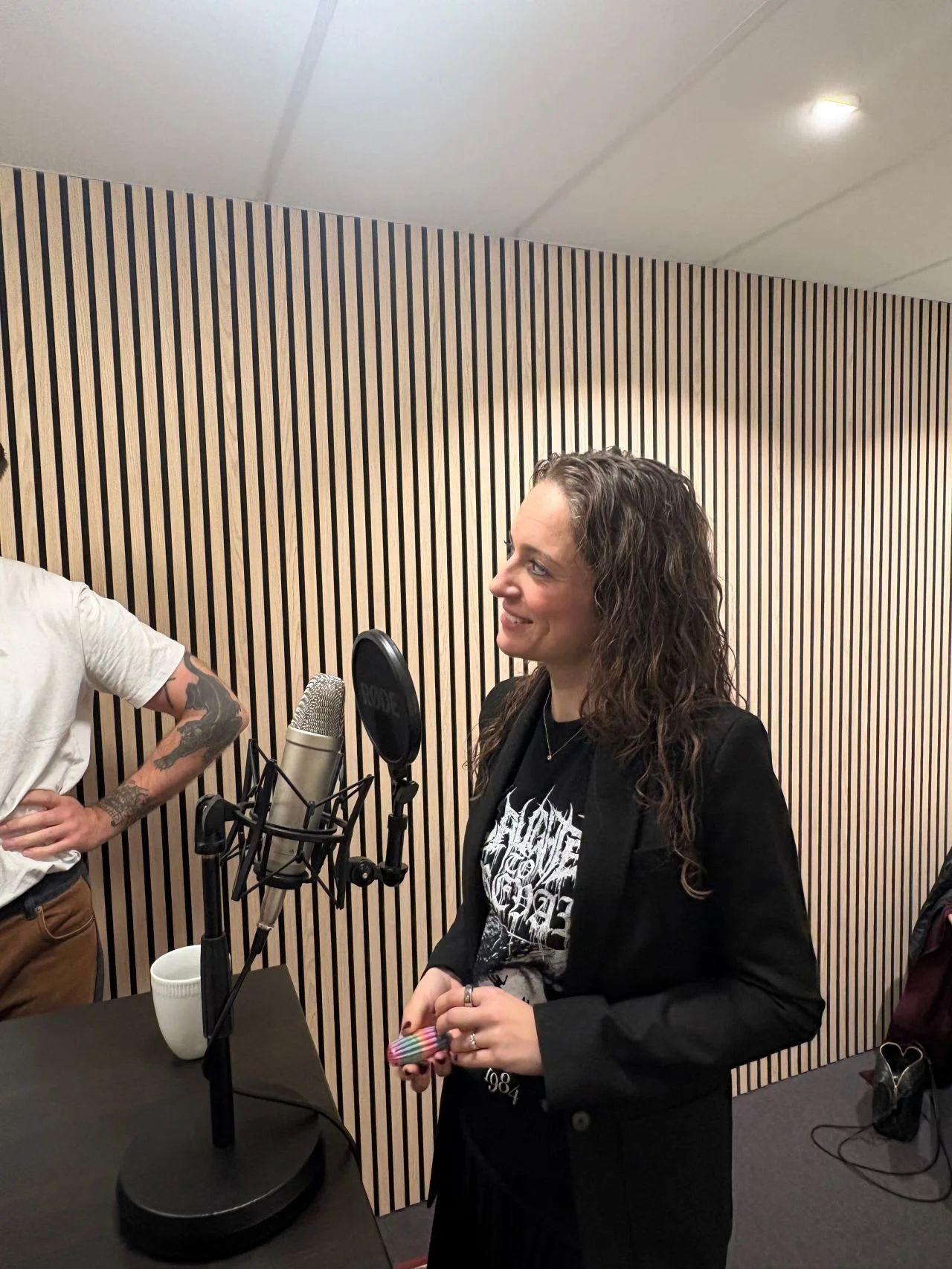 En kvinde med langt, krøllet hår smiler og taler i en mikrofon i et lydstudie med træpanel bagved.