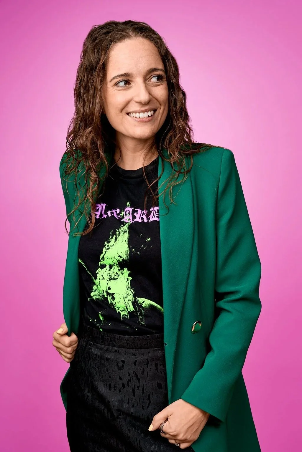 Kvinde med lange, vågede brune hår, iført en sort T-shirt med neongrøn grafik, en grøn blazer, står mod en lyserosa baggrund, smiler og ser til højre.