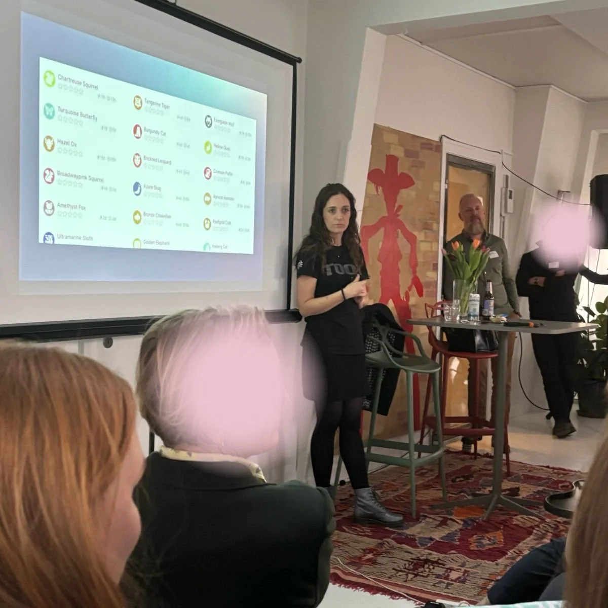 En kvinde står ved en præsentationsskærm foran en gruppe mennesker. Bag hende er der en mand med en skål med blomster og en person med blond hår. Der er nogle borde med drikkevarer og en grøn væg med et rødt symbol.