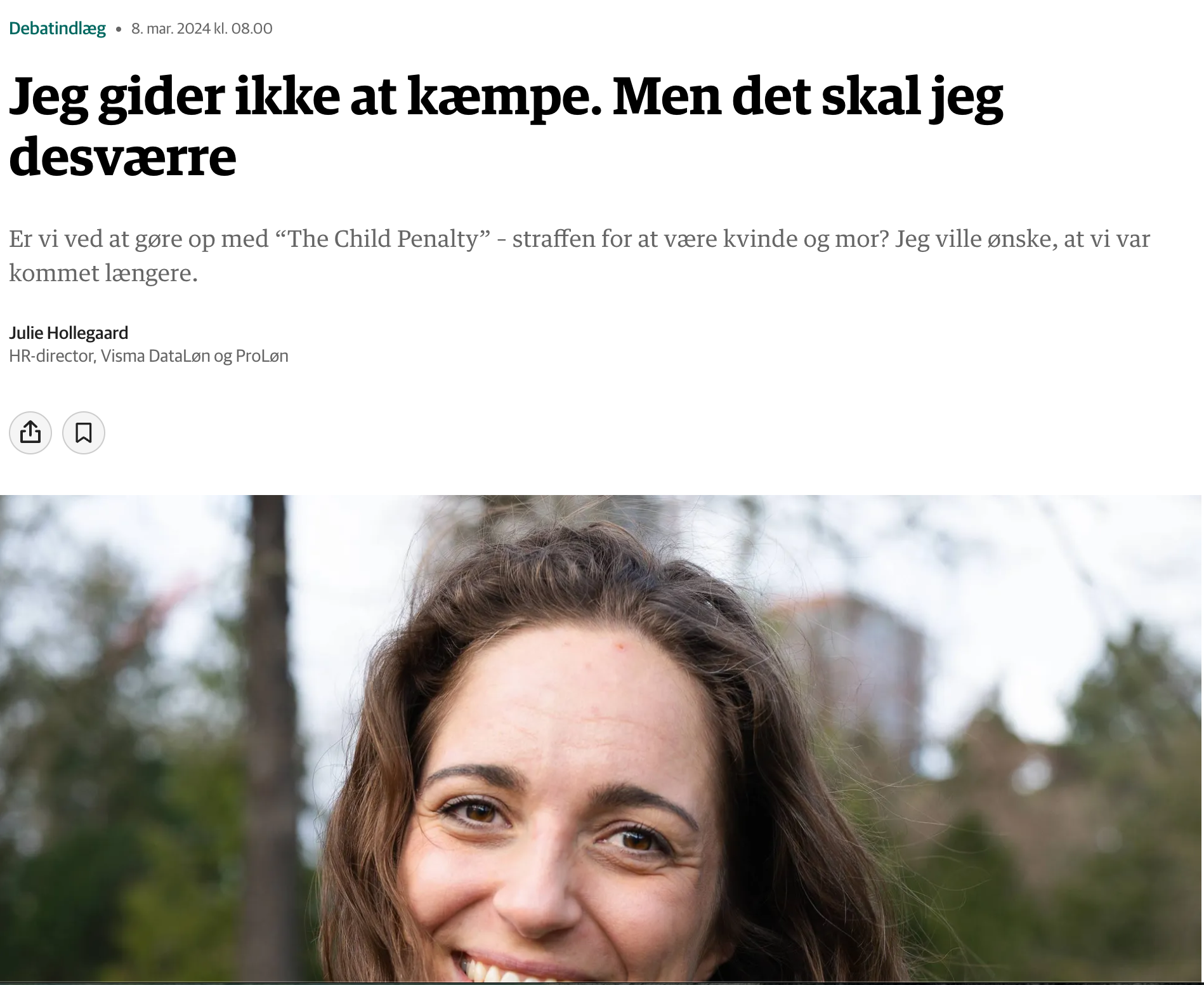 En kvinde med brunt, bølget hår smiler i et udendørs miljø med træer bagved. Det er en del af et portrætfoto, hvor hendes ansigt er i fokus.