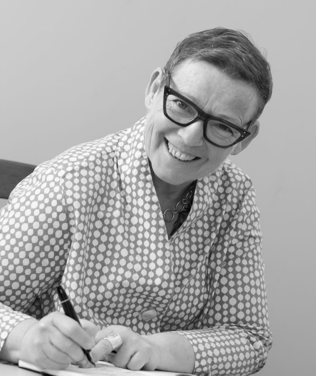 A woman with short hair, glasses, and a nose ring smiling while writing with a pen in a notebook.