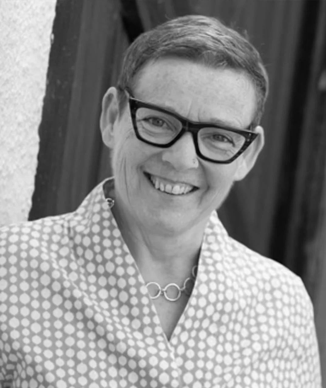 Black and white photo of a smiling woman with short hair, wearing glasses, a necklace, and a polka dot blouse, standing outdoors in front of a wooden background.