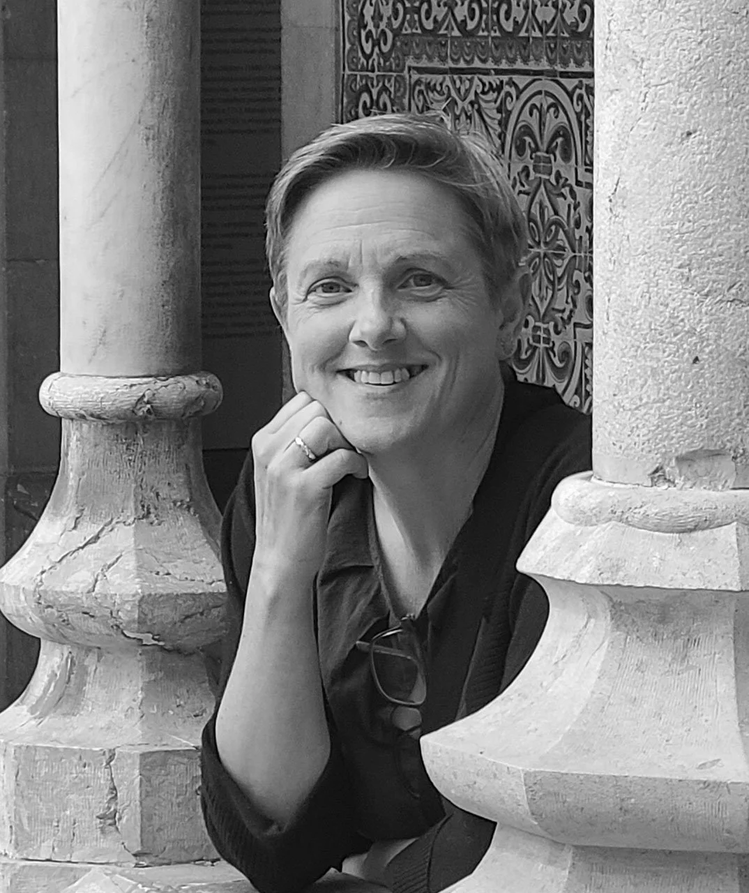 A smiling woman with short hair, wearing a dark top, resting her chin on her hand, framed by two stone columns with detailed decorative tiles in the background.