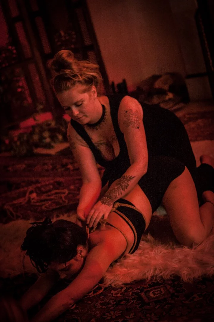 Two women in a dimly lit room; one is on all fours on a rug, and the other is kneeling behind her, holding her in a dominant position.