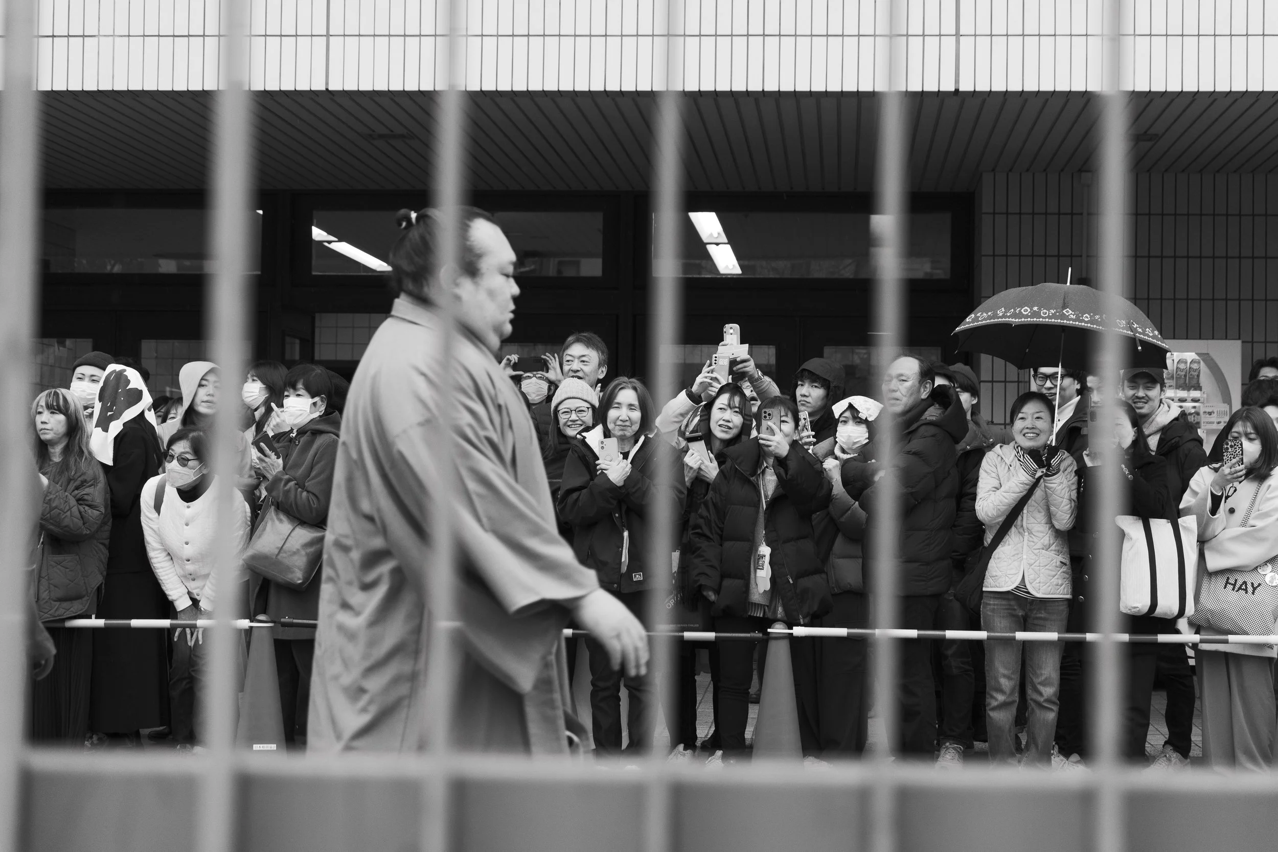 Black and white photo of a crowd of people standing outside a building, with some wearing masks and holding umbrellas, seen through metal bars.