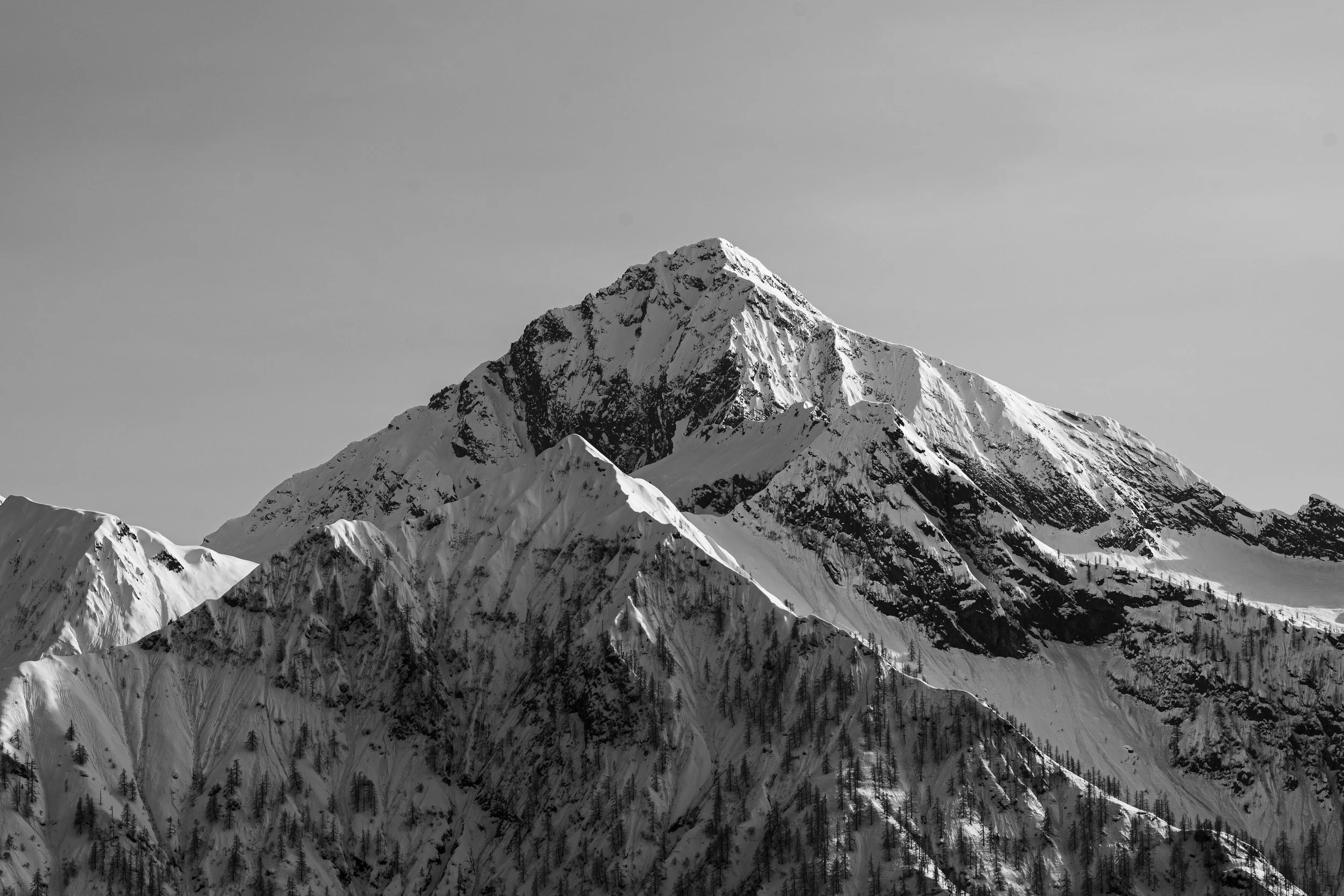Black and white photo of a  snow-covered mountain with a sharp peak, surrounded by smaller snow-covered peaks and sparse trees at the base.