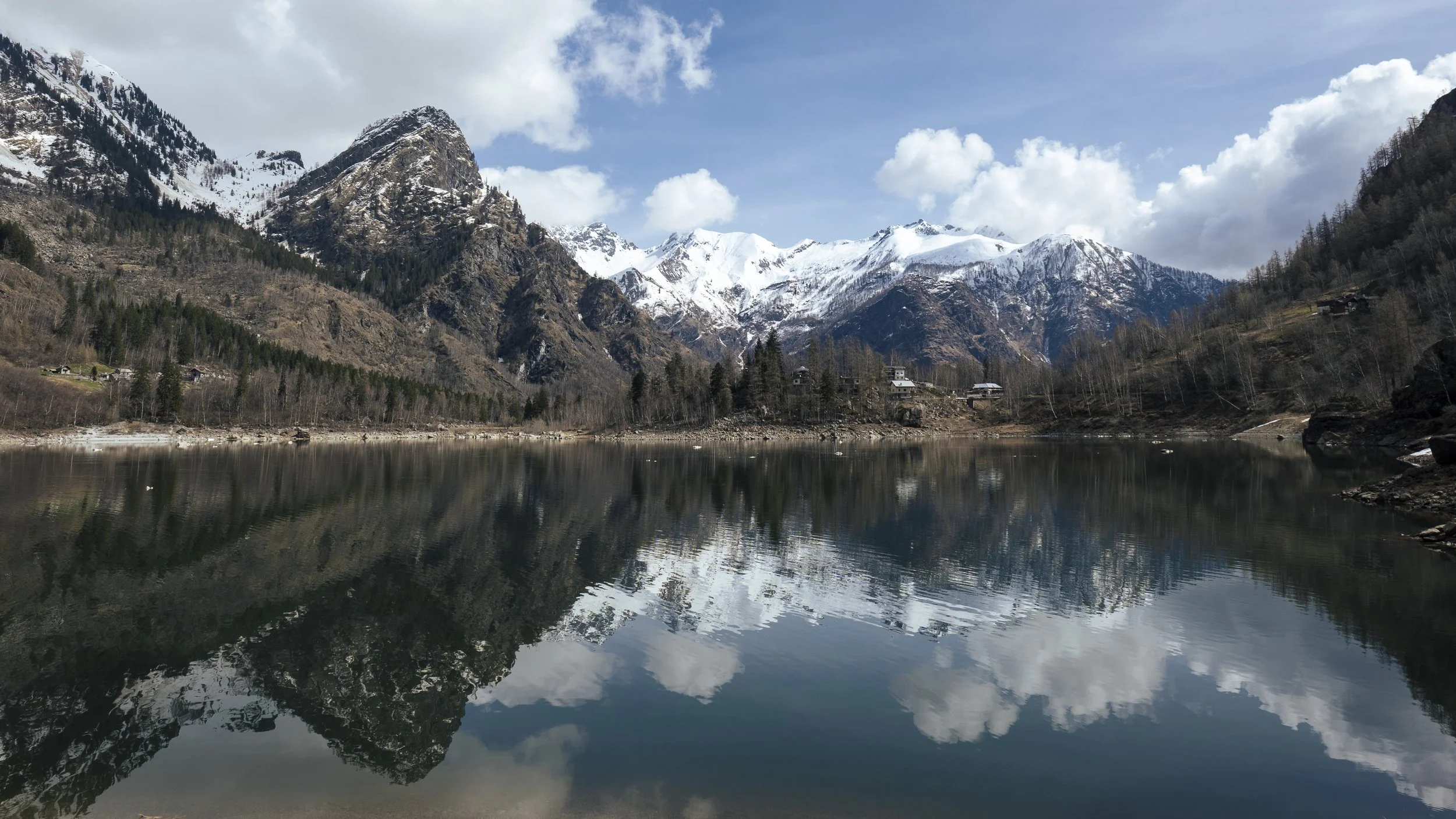 Snow-capped mountains reflected in a calm lake with a few small houses on the shoreline, surrounded by forest.
