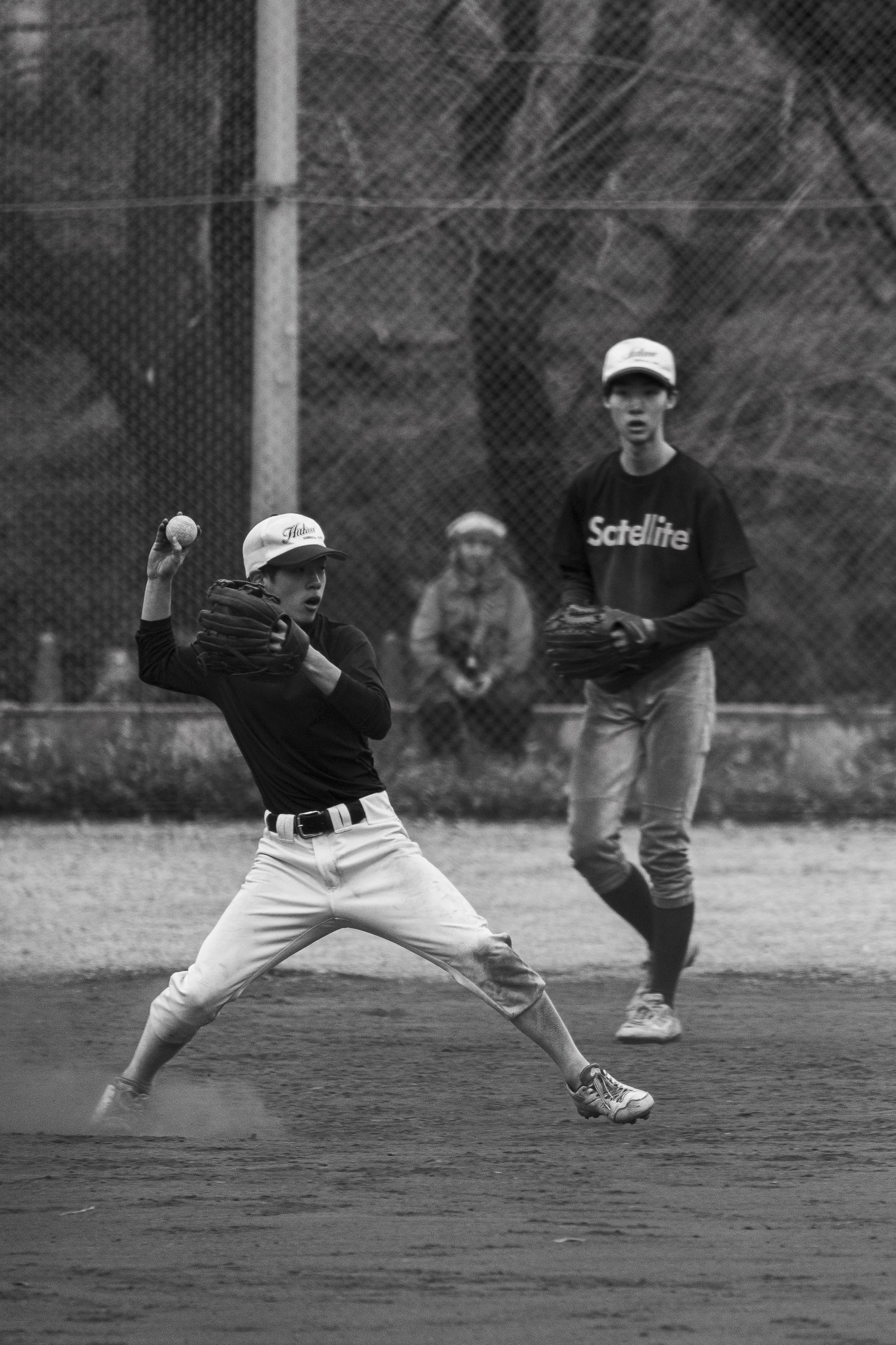 A black-and-white photograph of two young baseball players on a field during a game. One player is in a pitching stance, holding a baseball in his right hand and wearing a baseball glove on his left hand. The other player, standing nearby, is in a re