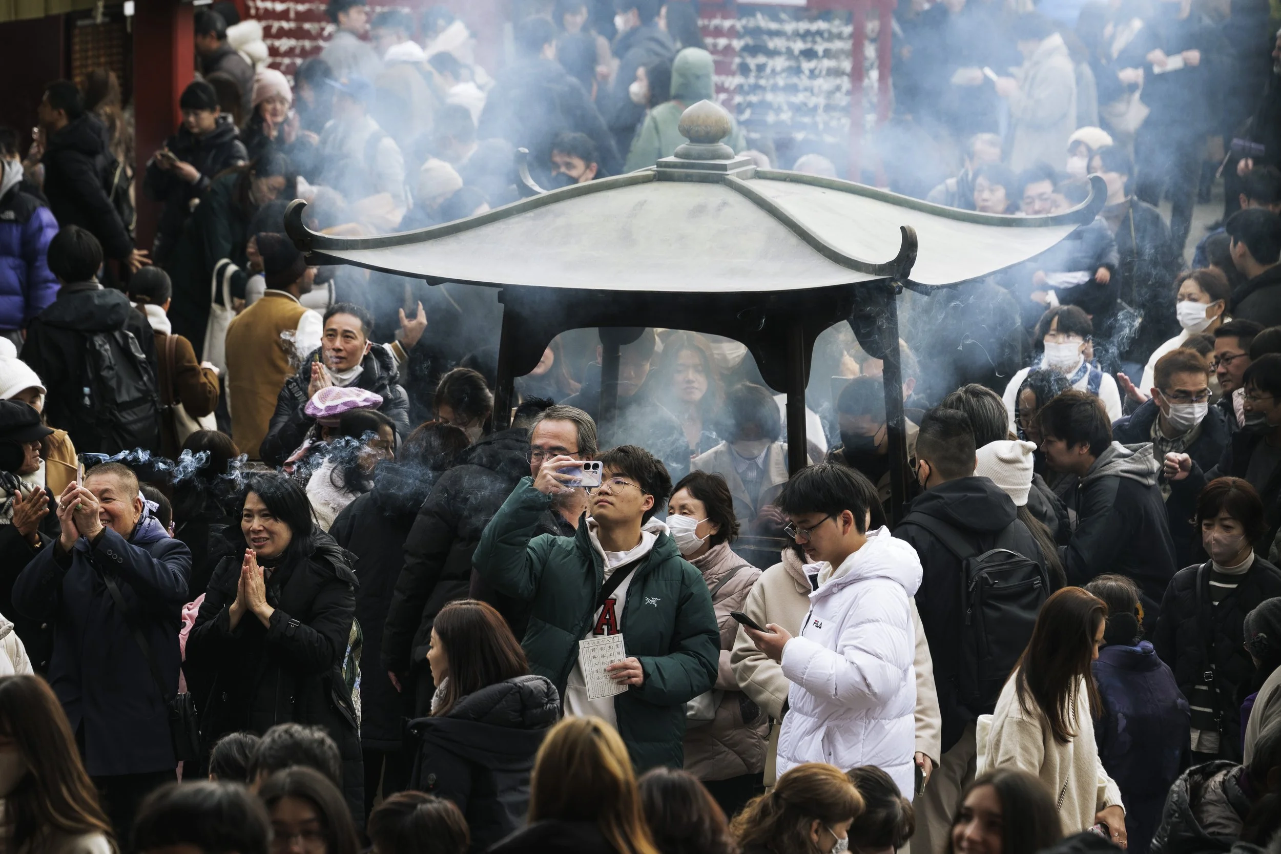 A large crowd of people gathered outdoors, many wearing masks, with some praying and others taking photos. There is a traditional Asian-style pavilion in the center emitting smoke.