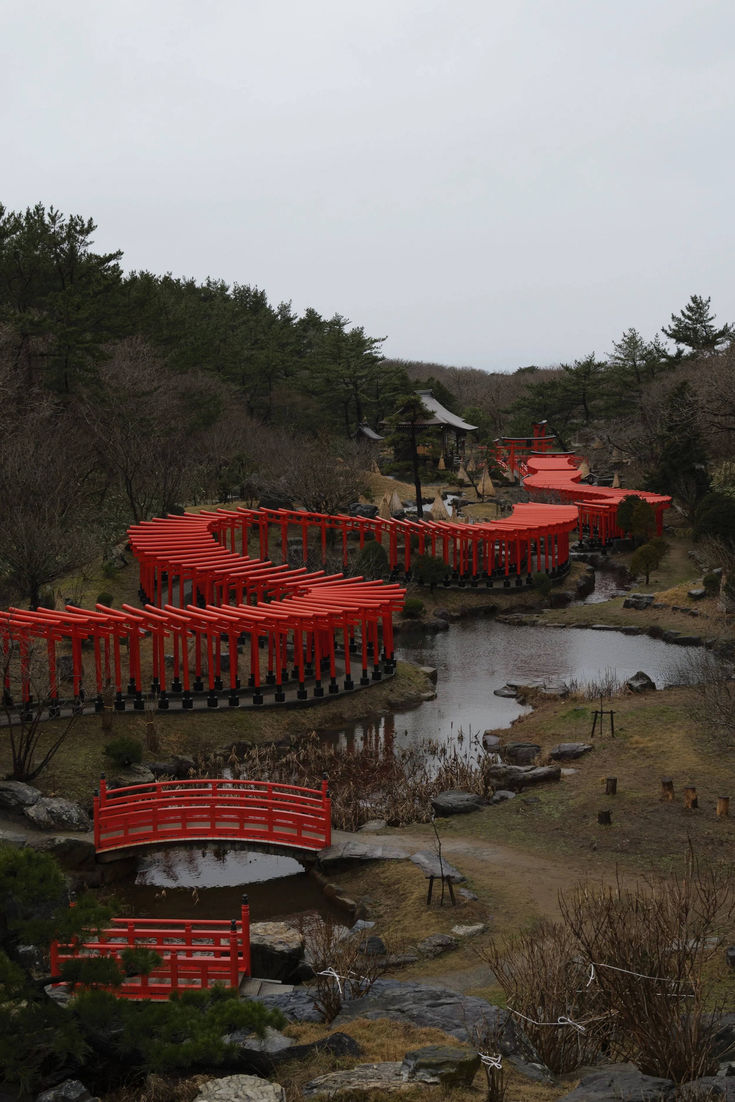 A traditional Japanese garden featuring a winding red torii gate pathway over a small pond, surrounded by trees and shrubs, with a small temple structure in the background under an overcast sky.