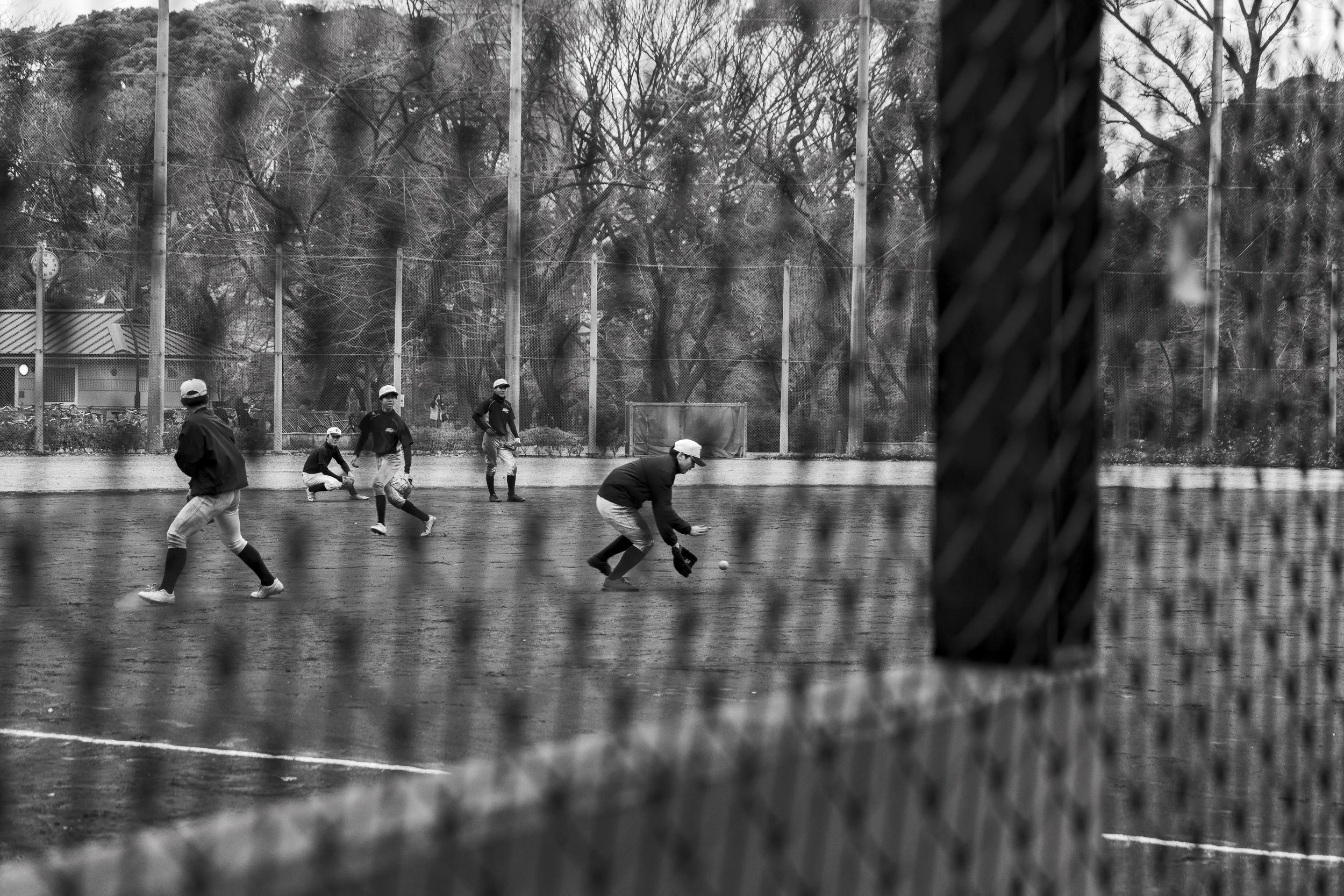 Black and white photo of children playing baseball on a field, viewed through a chain-link fence.